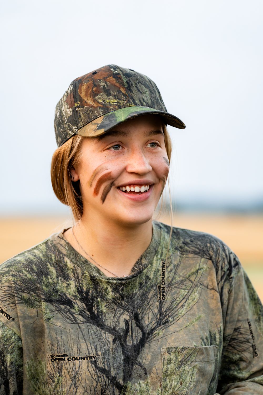A person wearing camouflage clothing and baseball cap smiles while outdoors.