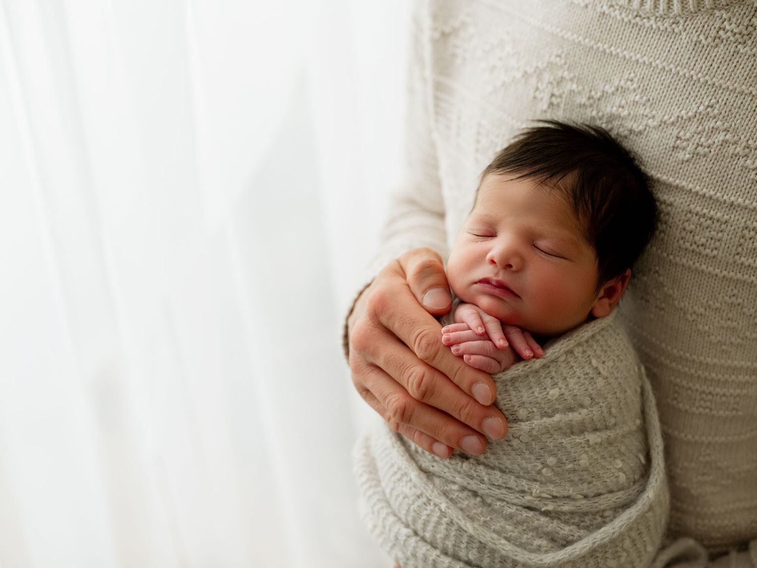 A sleeping newborn baby wrapped in a soft white blanket with gentle natural light streaming in from the side.