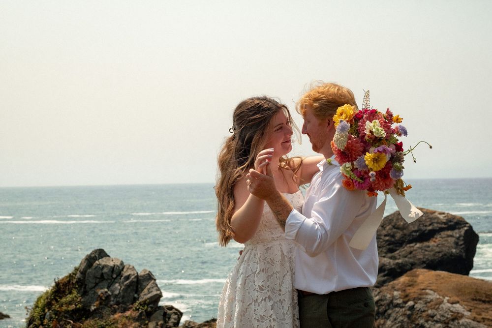 Coastal wedding photography with couple embracing against dramatic ocean backdrop and colorful flowers.