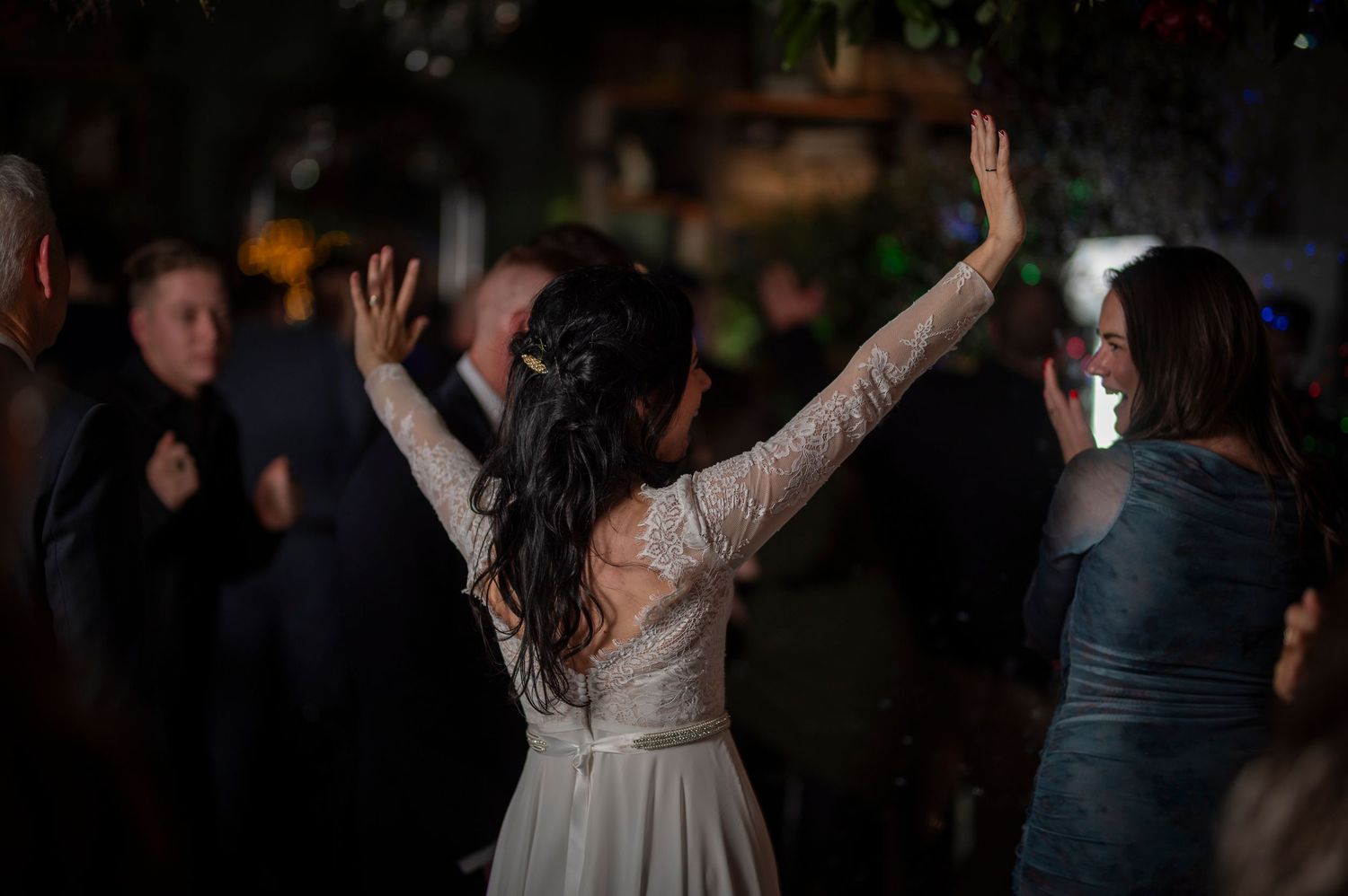 A person in a white lace wedding dress dances with raised arms at an evening reception celebration.