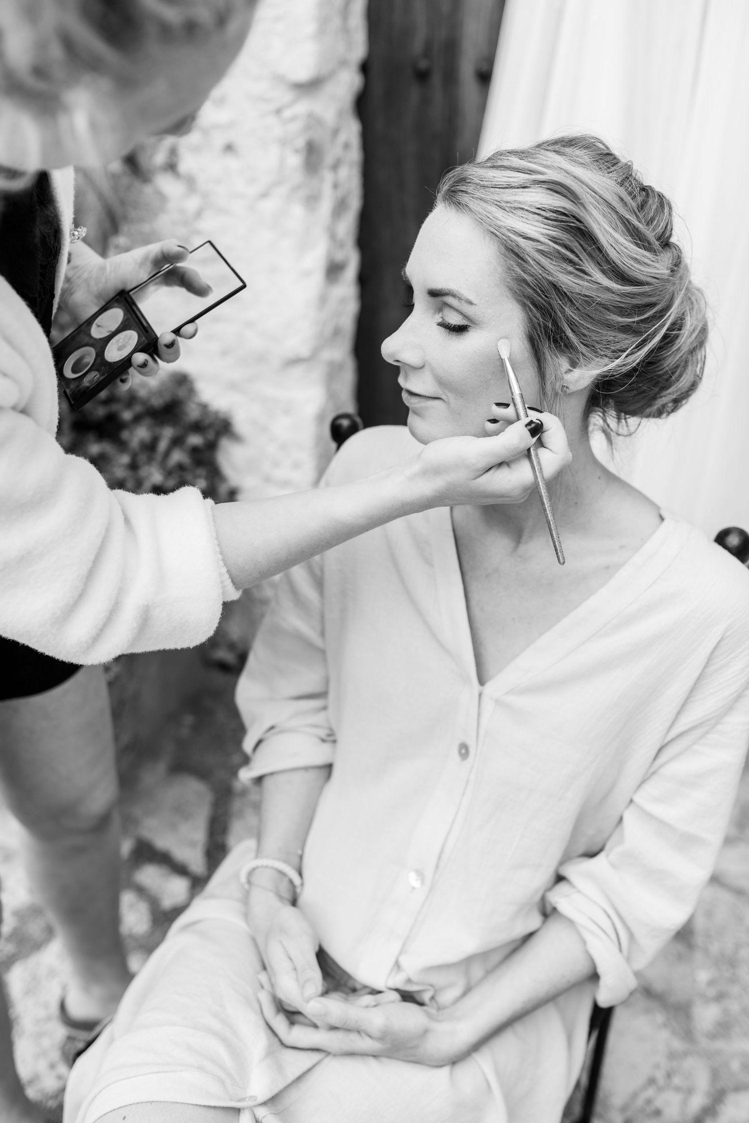 Schwarz-Weiß-Close-up einer Visagistin, die einer Braut, die ein weißes Kleid trägt, beim Getting Ready auf Mallorca Kosmetika aufträgt.