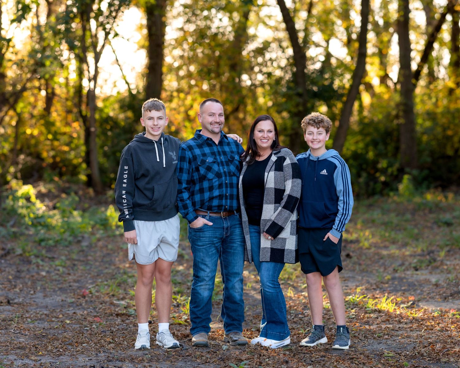 Family of four smiles together outdoors during a fall portrait session in a wooded setting.