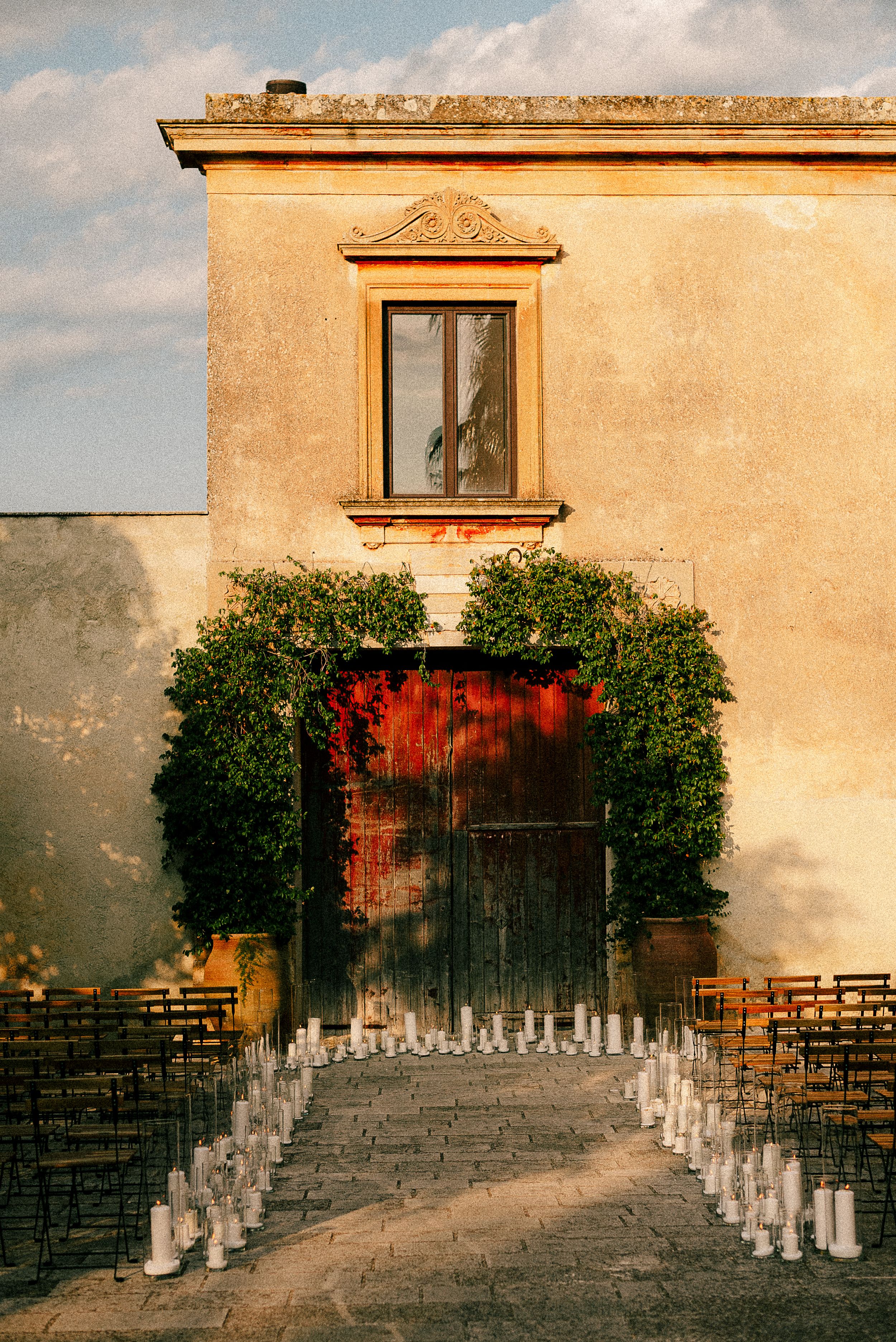 Wedding ceremony setup with white chairs arranged in curved rows facing a rustic doorway adorned with greenery and red accents.
