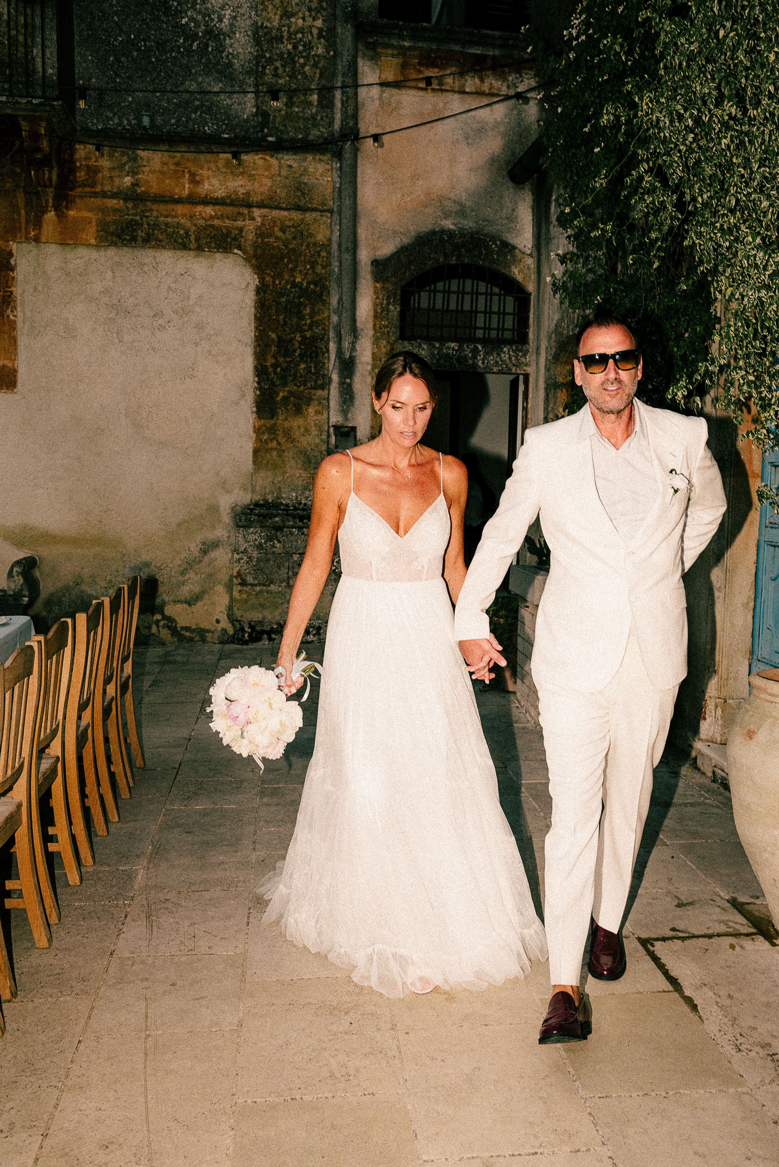 Newlyweds in white attire walk through a rustic venue with wooden chairs.