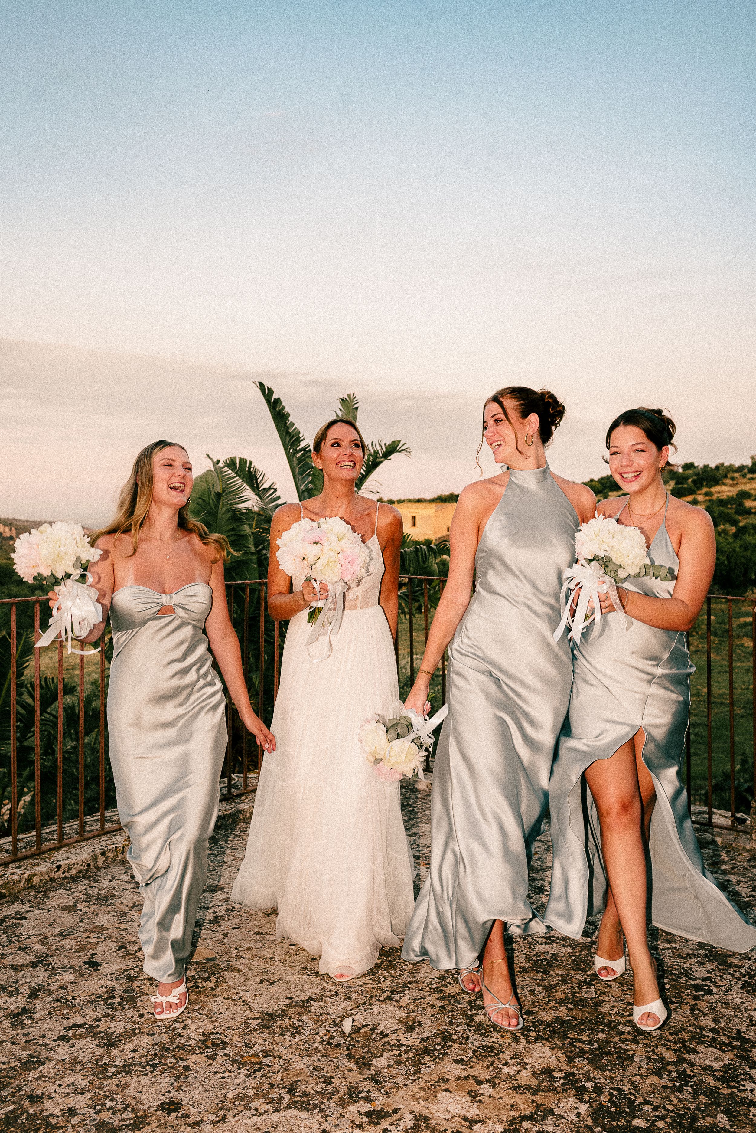 Bridal party in silver dresses and white gown share candid laughing moments during sunset photoshoot on outdoor terrace.