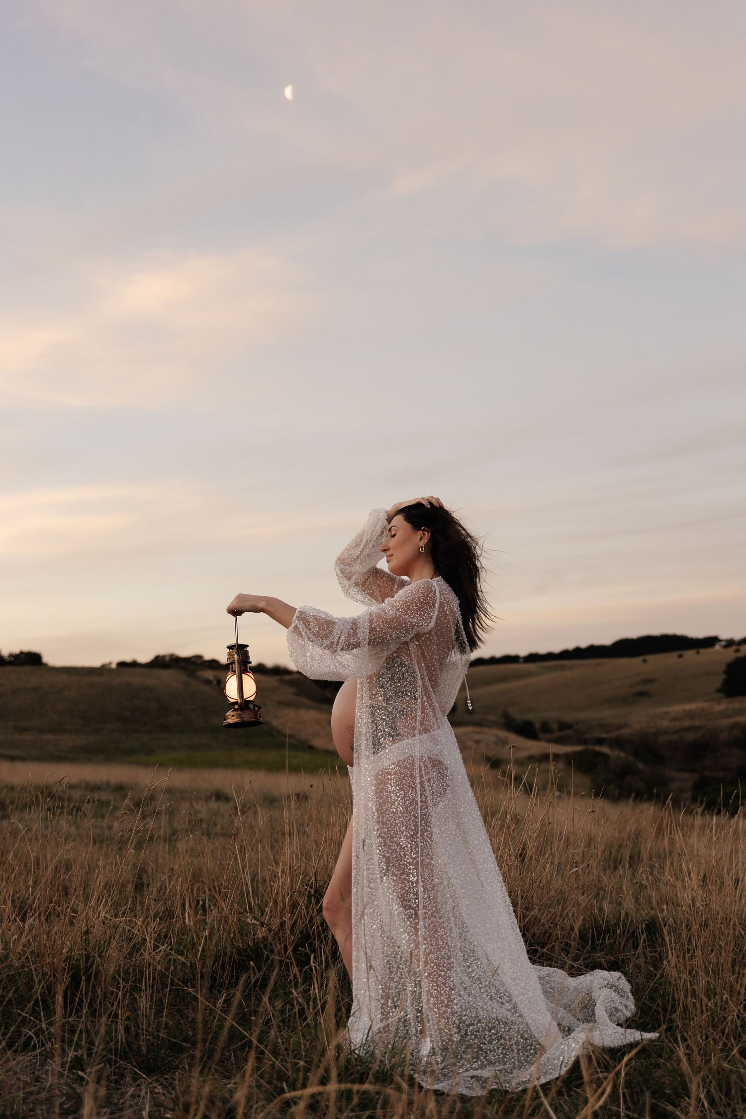 Figure in flowing lace dress holds lantern at dusk in rustic field with warm sunset sky and rolling hills.