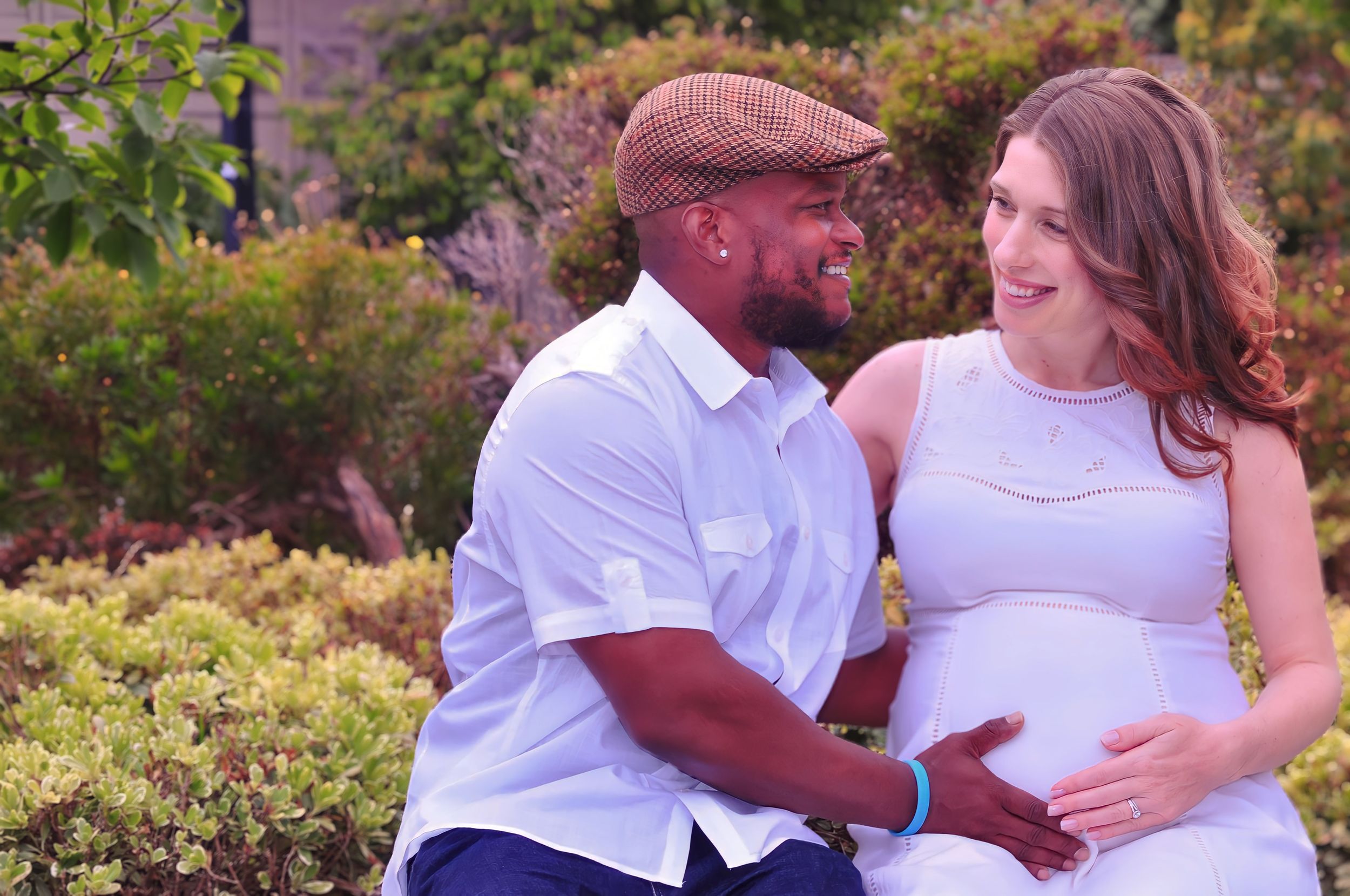 Expectant couple smiles lovingly at each other while sitting in a garden during a maternity photoshoot.