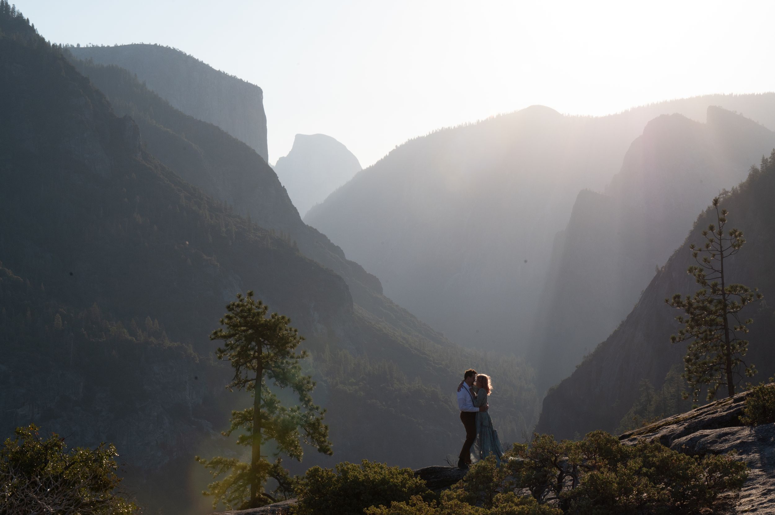 Silhouetted figure stands on mountain ledge overlooking misty Yosemite Valley at sunrise with sun rays streaming through.