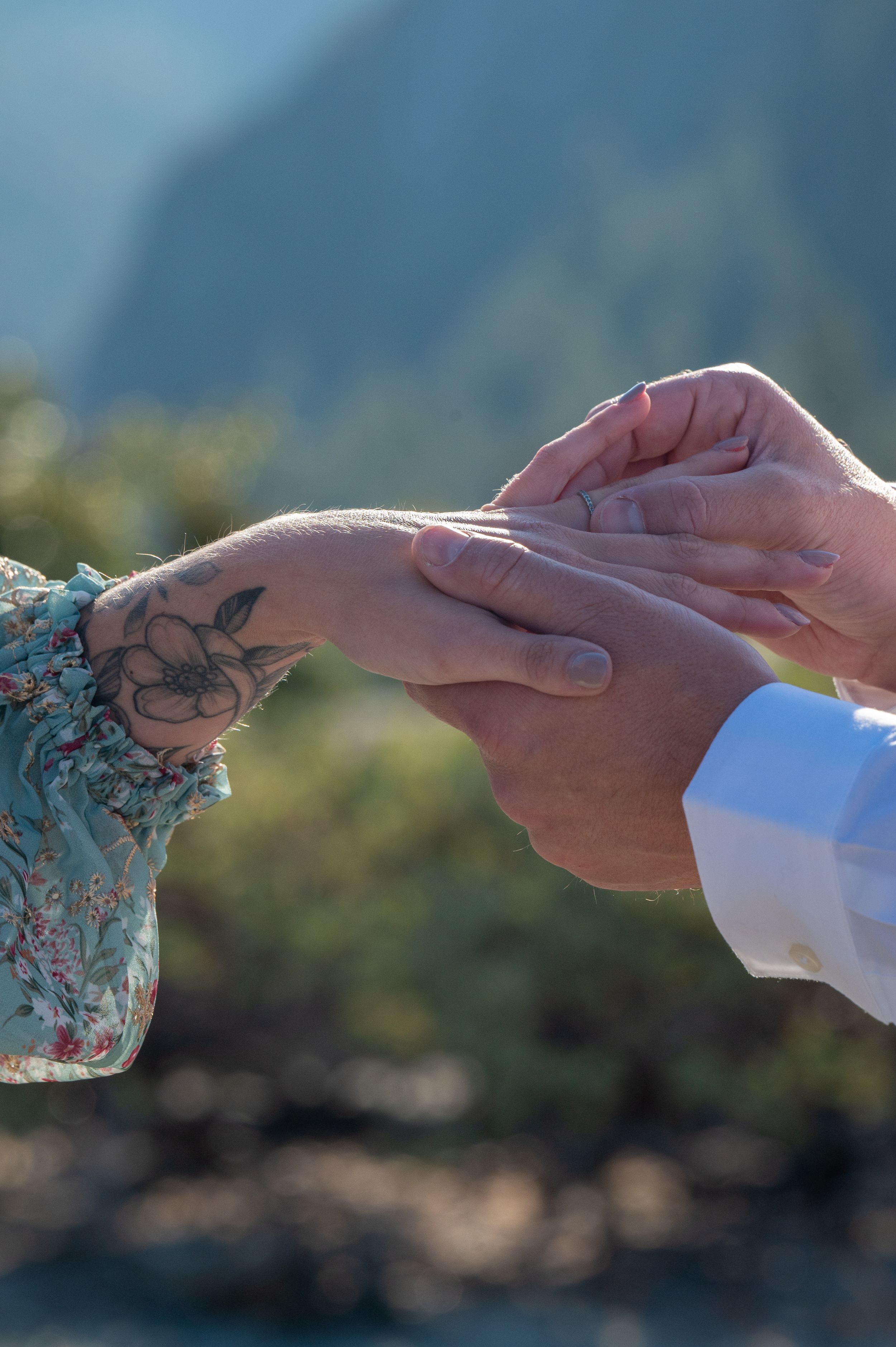 Close-up view of hands exchanging rings during an outdoor ceremony with a floral tattoo visible on one person's wrist.