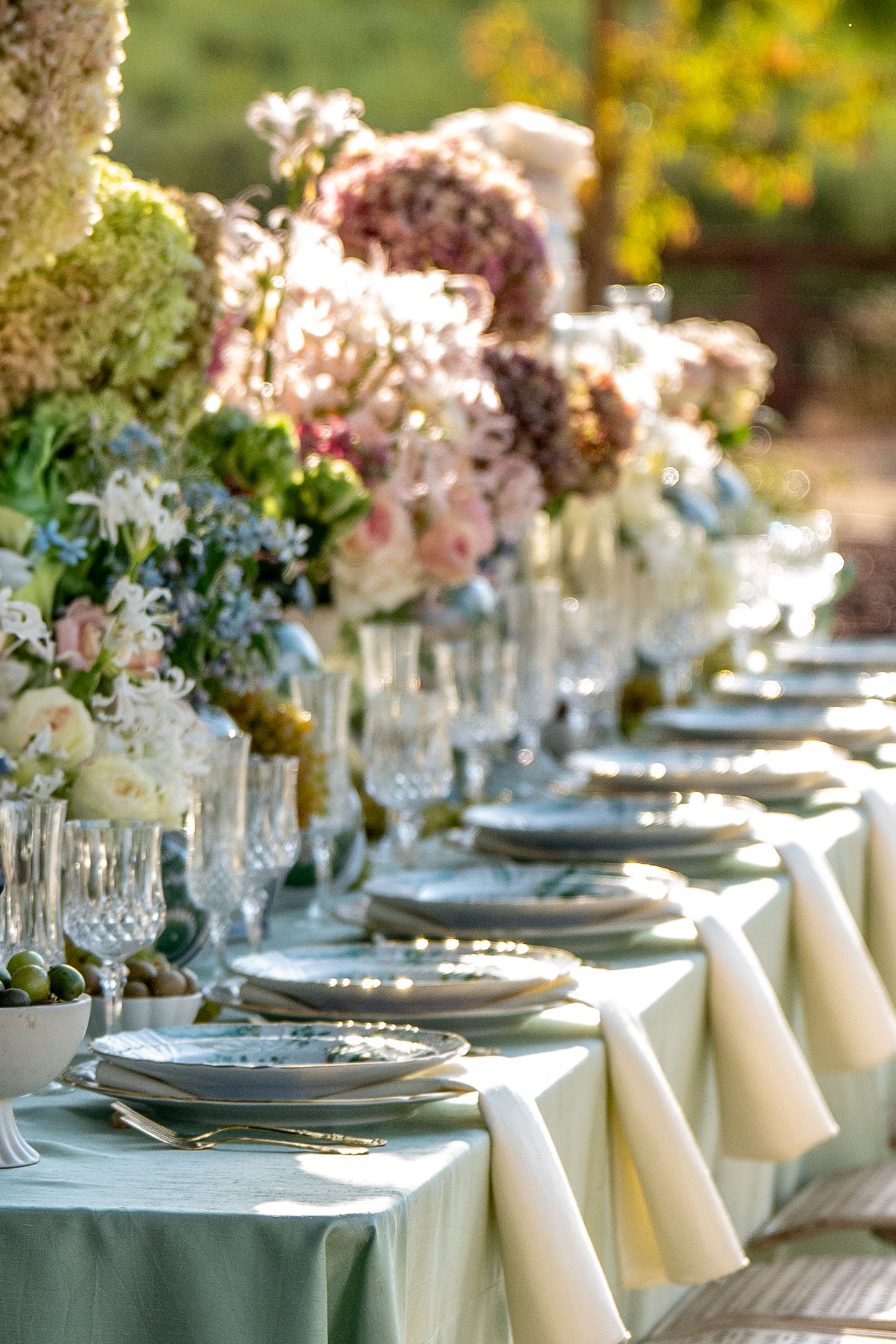 Elegant table setting with crystal glassware and pink floral centerpieces on sage green and cream linens.