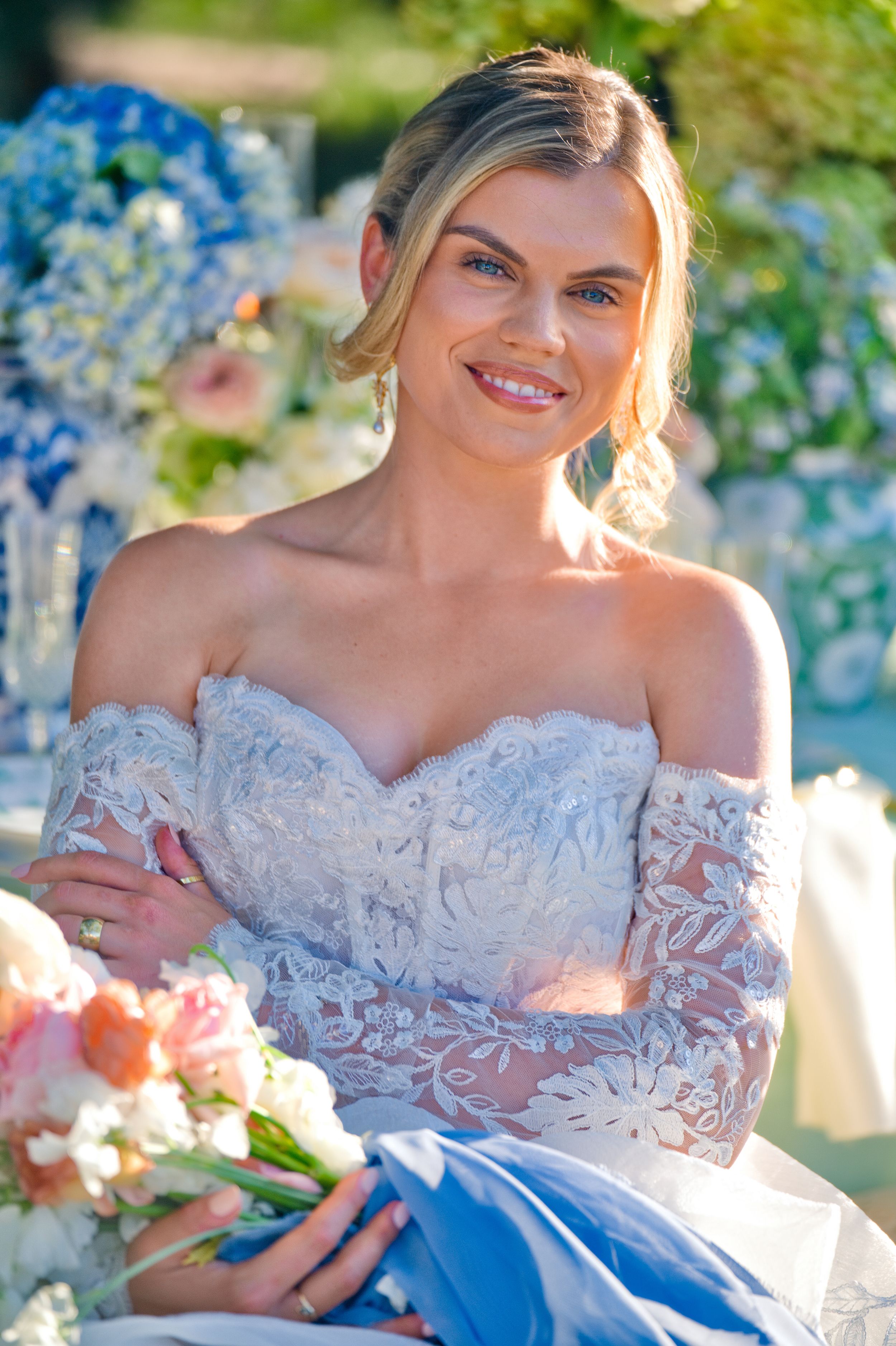 Beautiful bride in off-shoulder white lace gown poses outdoors with blue hydrangeas and pink flowers in background.