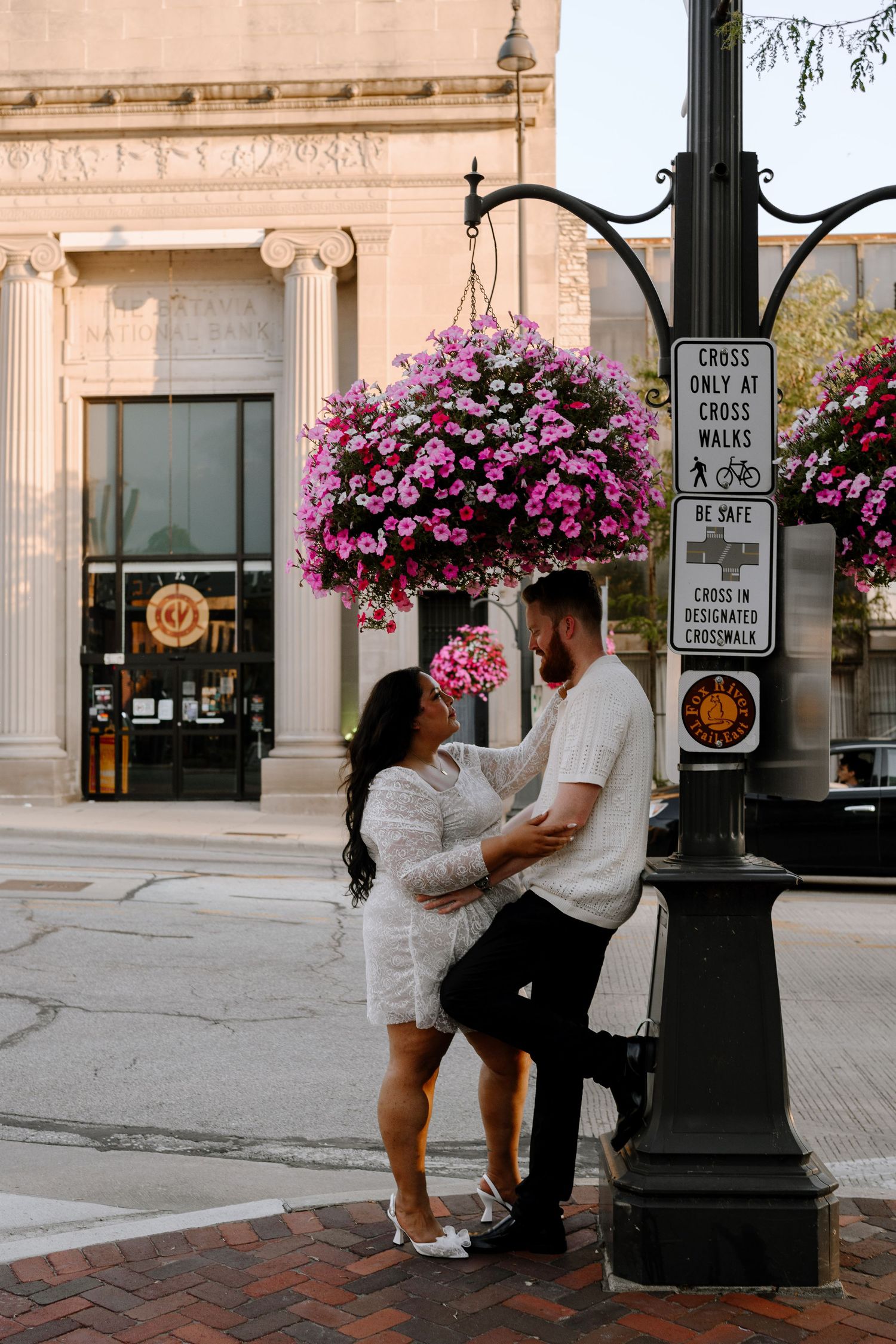 A romantic couple embraces near a vintage lamp post with vibrant pink hanging flower baskets on a city street.