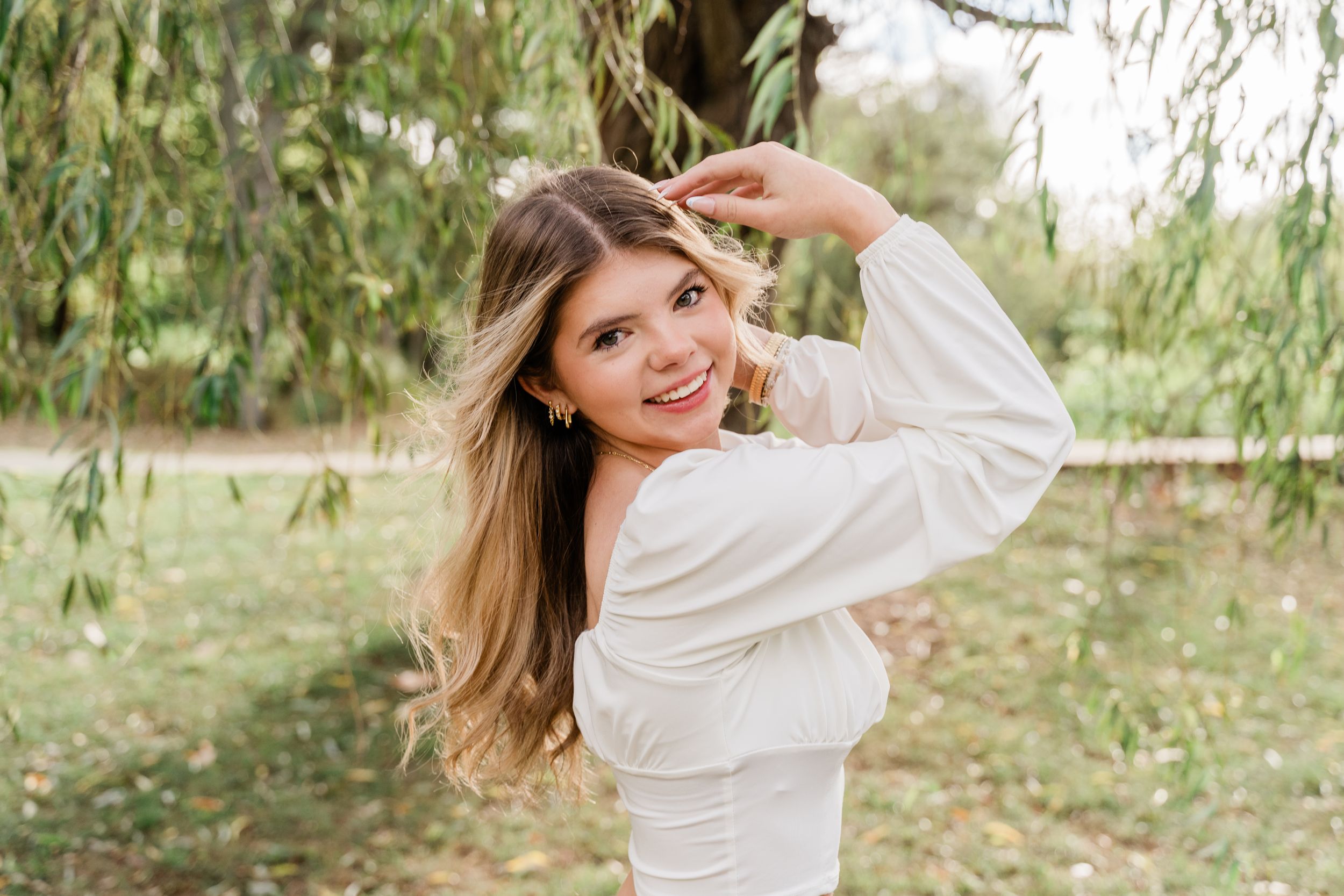 A woman in a white sweater poses playfully under a willow tree on an autumn day.