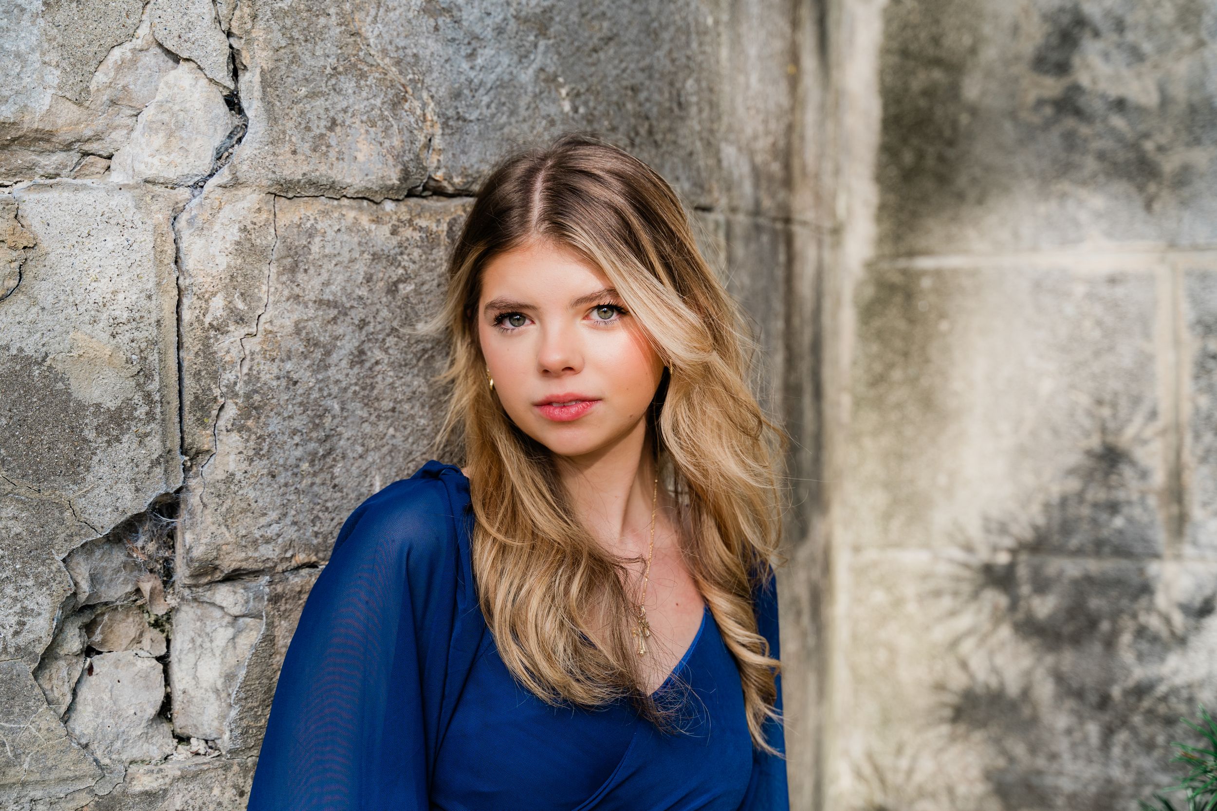 A woman in a blue sweater poses against a textured stone wall as natural light illuminates her face.