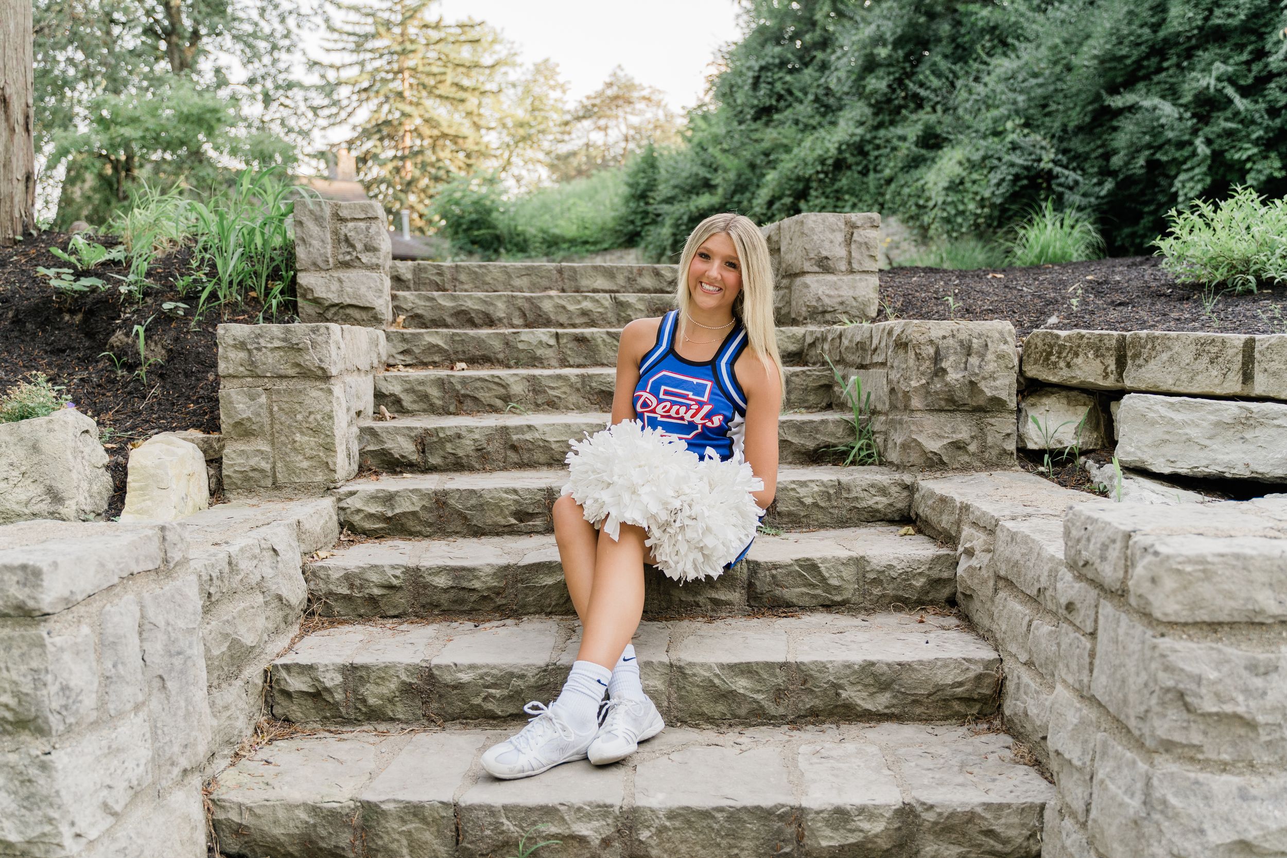 A cheerleader in a blue uniform sits on stone steps outdoors surrounded by green foliage.
