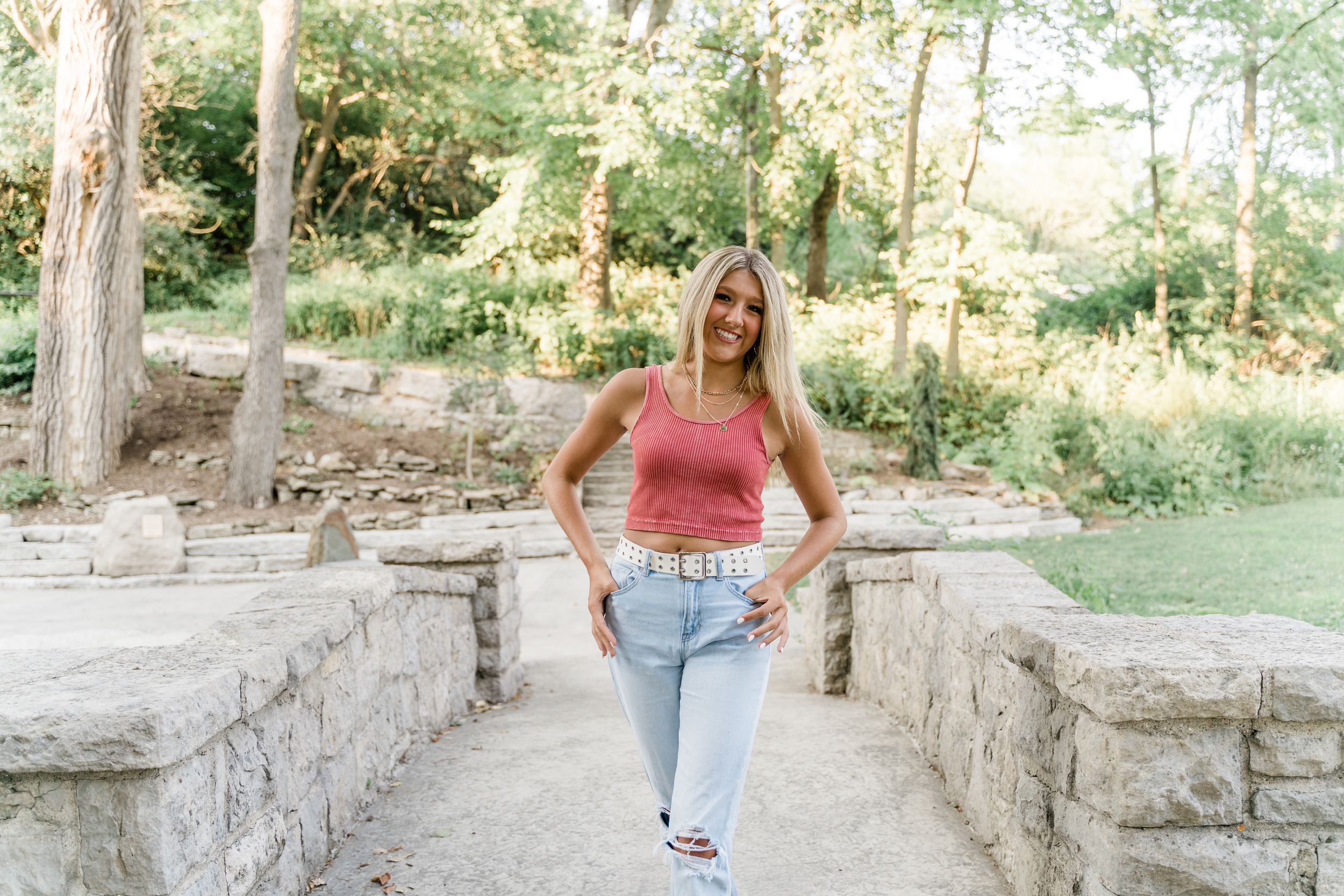 A person in light colored clothing poses on stone steps surrounded by green trees and natural scenery.