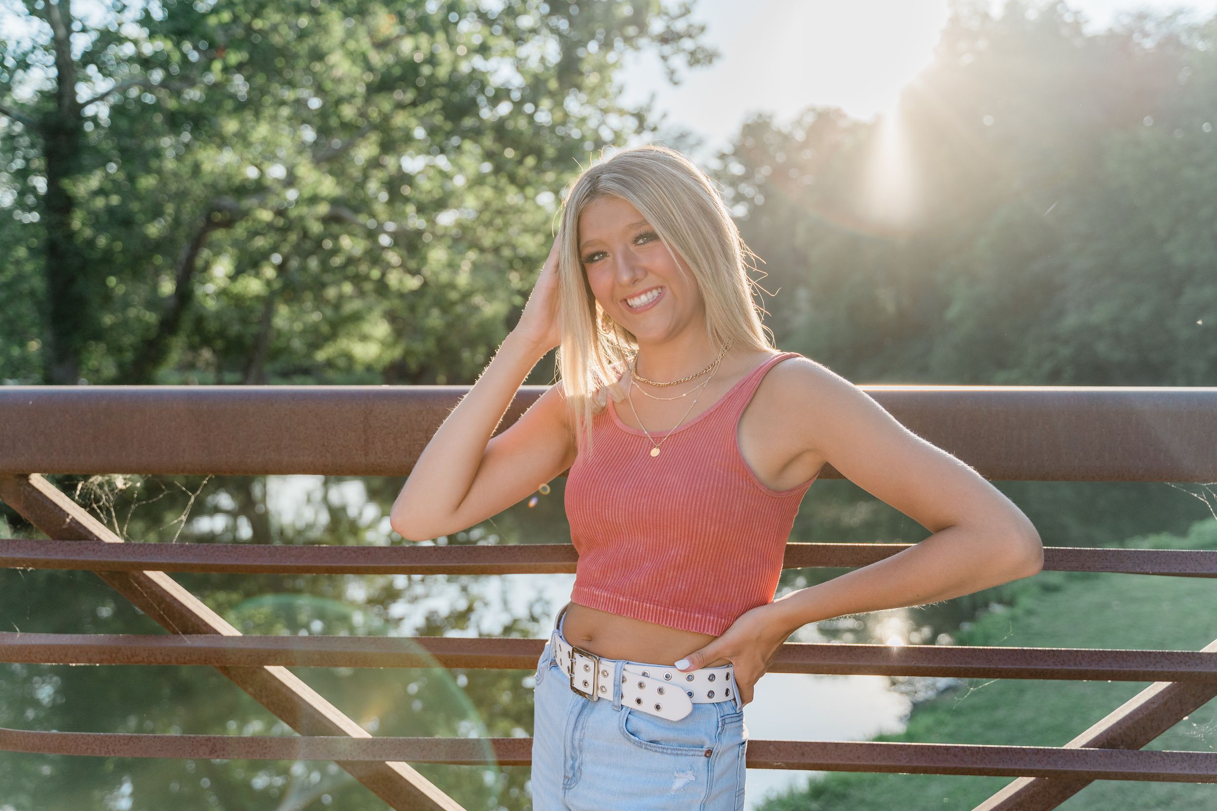 Young woman in coral tank top and denim shorts sitting outdoors on a park bench during golden hour.