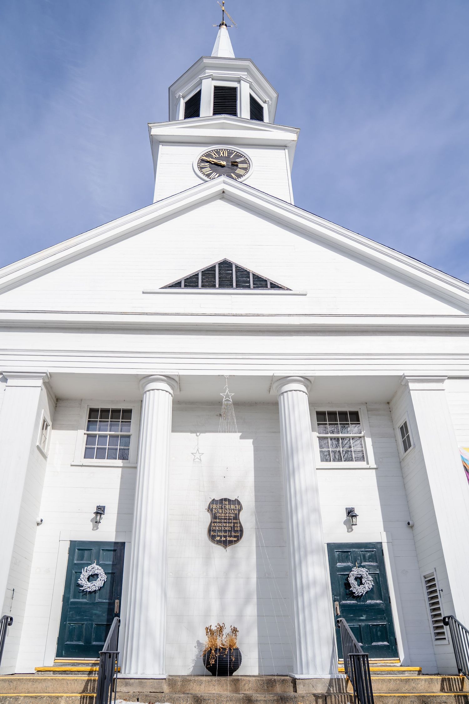 First Church of Wenham with a tall steeple, double green doors and decorative holiday wreaths against blue sky. First Church of Wenham