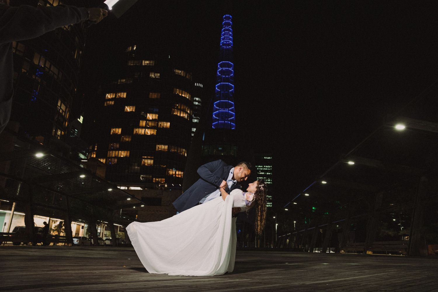 A couple in wedding attire dances together in front of illuminated city buildings at night.