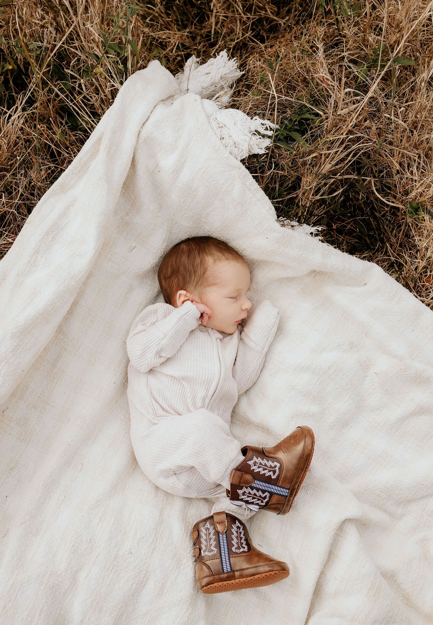 Sleeping baby on white blanket wearing brown leather boots in dried grass setting.