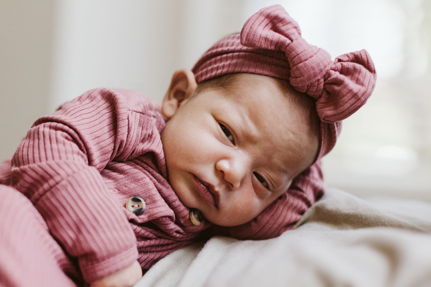 Newborn baby in pink ribbed outfit with bow lying on soft surface.