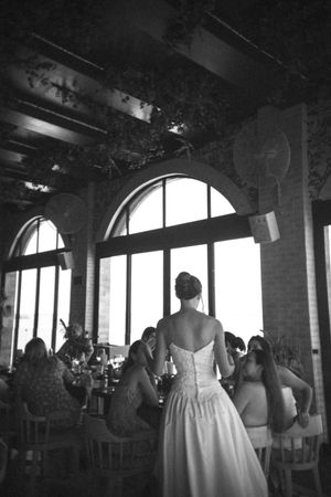 A black and white photograph of a bride in a strapless gown standing in front of arched windows at a reception venue.