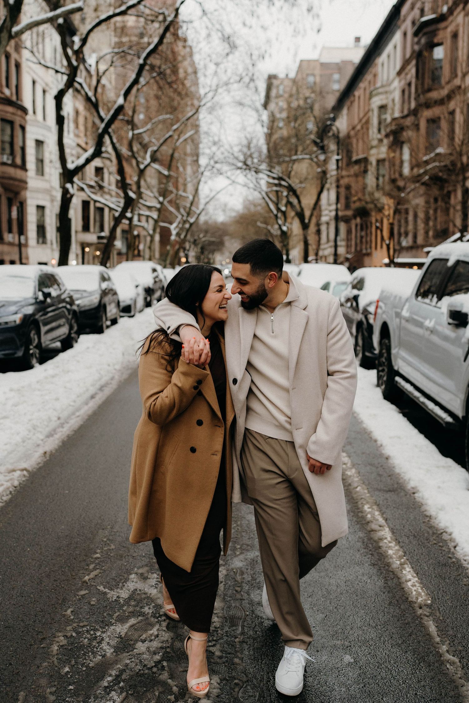 Couple laughing together while walking down snow-covered urban street in winter coats.