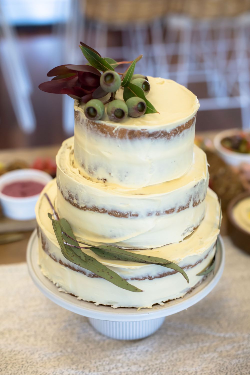A rustic semi-naked white tiered cake decorated with fresh eucalyptus leaves and dark berries on a white pedestal stand.