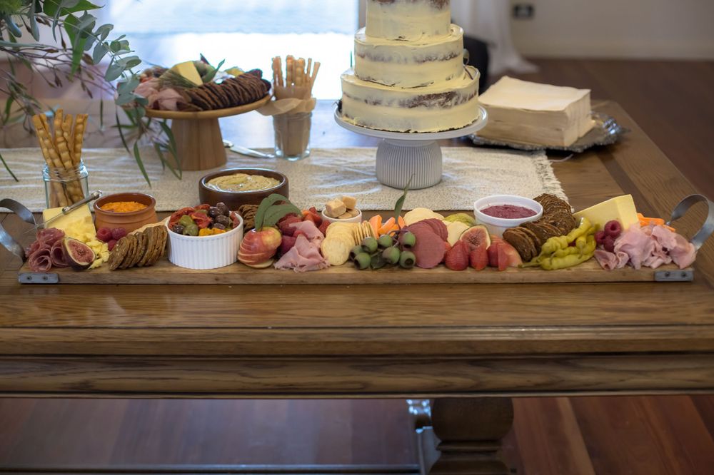 A rustic wooden charcuterie board displays meats, cheeses, fruits and breads alongside a tiered white cake on a stand.