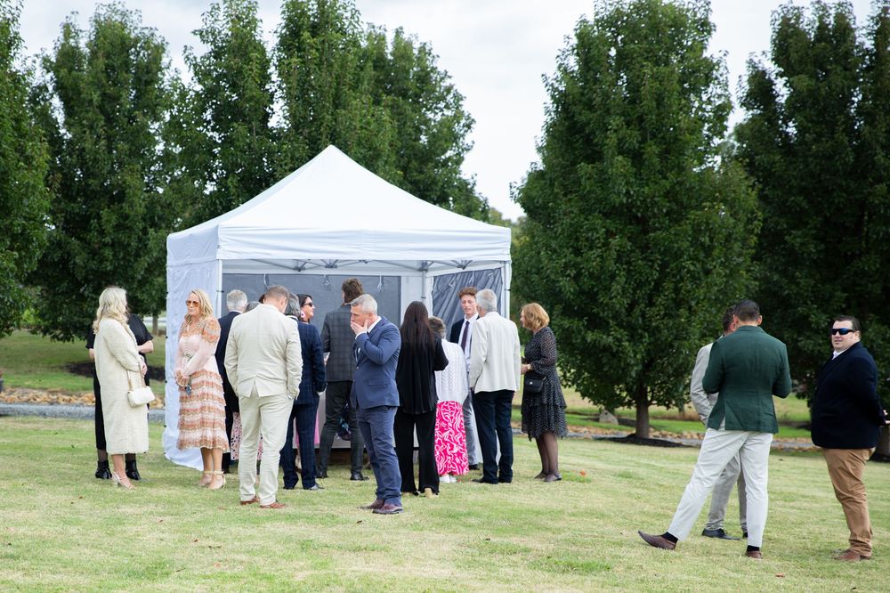 A group of people gather under a white pop-up tent on a grassy lawn surrounded by tall green trees.