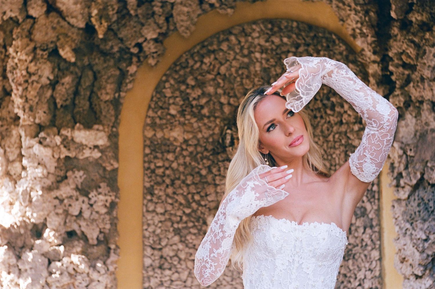 Elegant white lace wedding dress with long sleeves shown against textured stone wall with arched architectural detail.