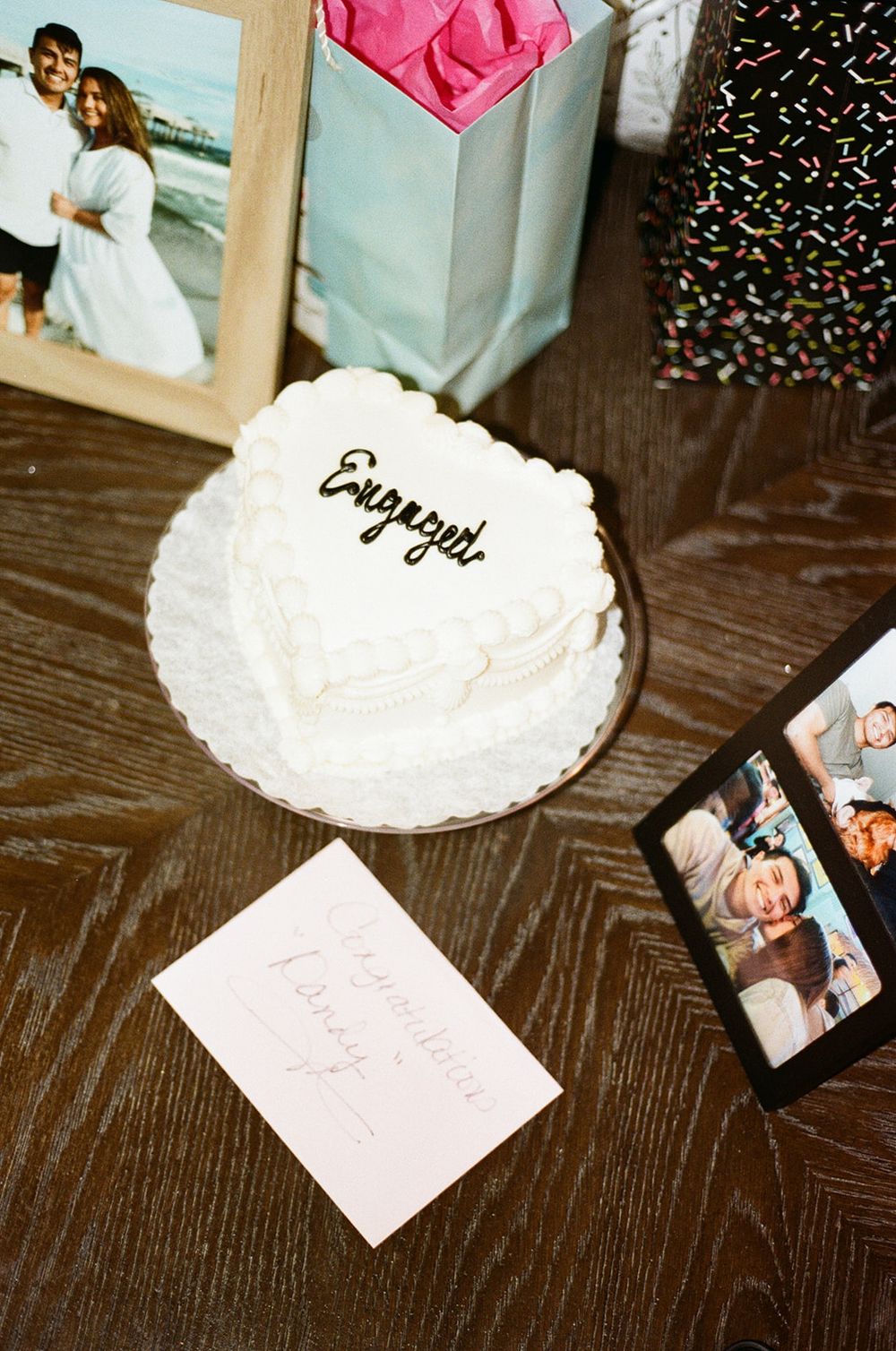 A white celebration cake with "Enjoy You" text sits on a dark wooden table with photos and a gift bag nearby.