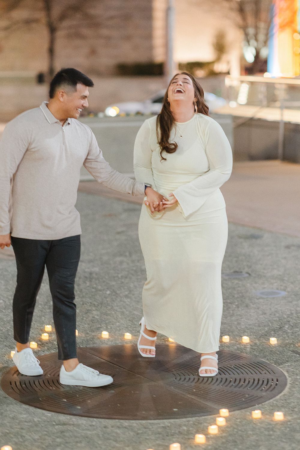 A couple in white and grey outfits dance together in a romantic setting surrounded by glowing candlelight.
