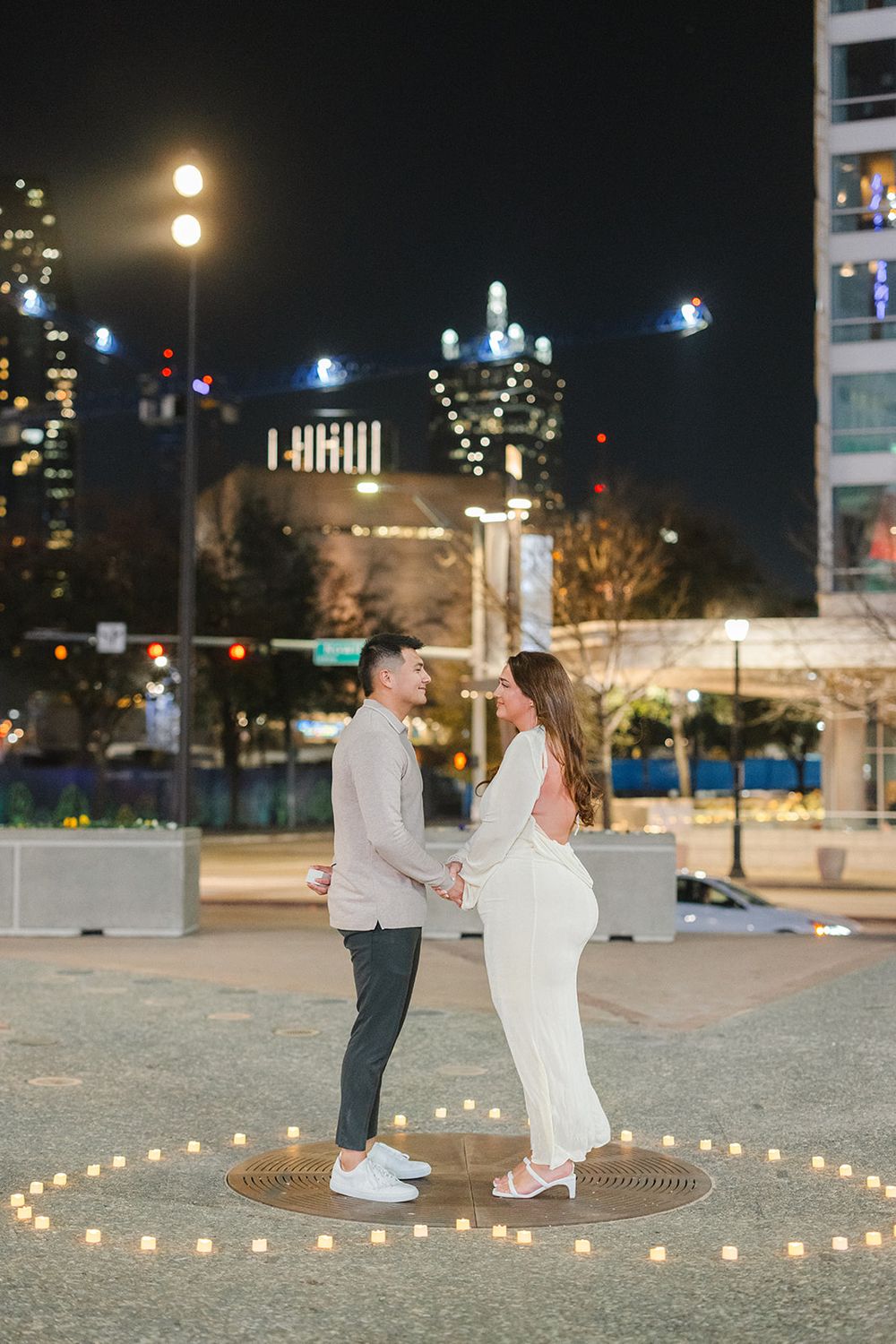 A couple stands together in the center of a heart made from tea lights with a city skyline glowing in the background.