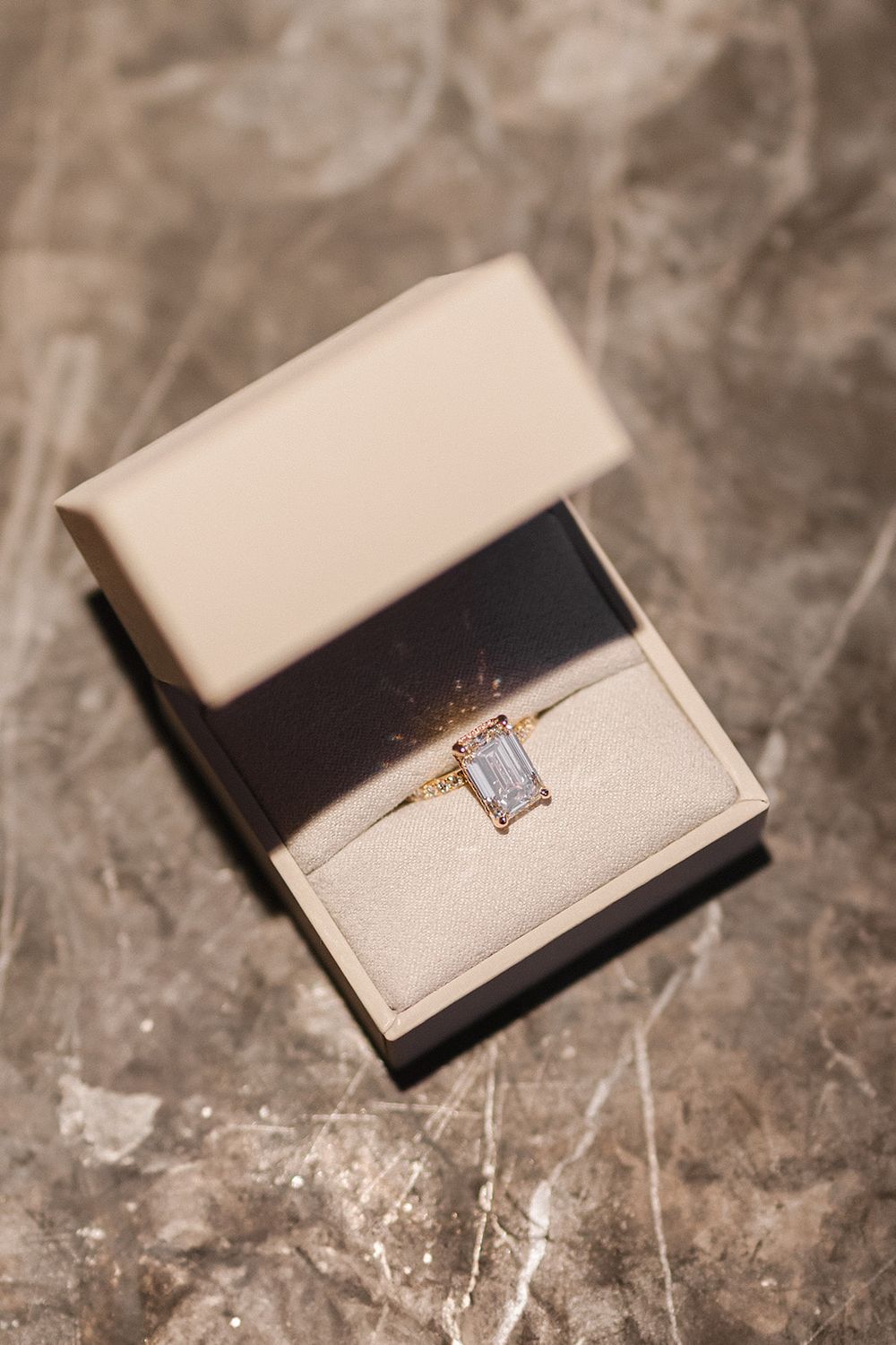 A delicate gold ring with an emerald-cut diamond displayed in an elegant cream-colored jewelry box on marble surface.
