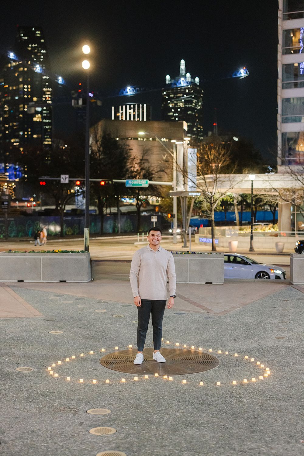 A person stands in the center of a heart made from string lights at night with city buildings illuminated in the background.