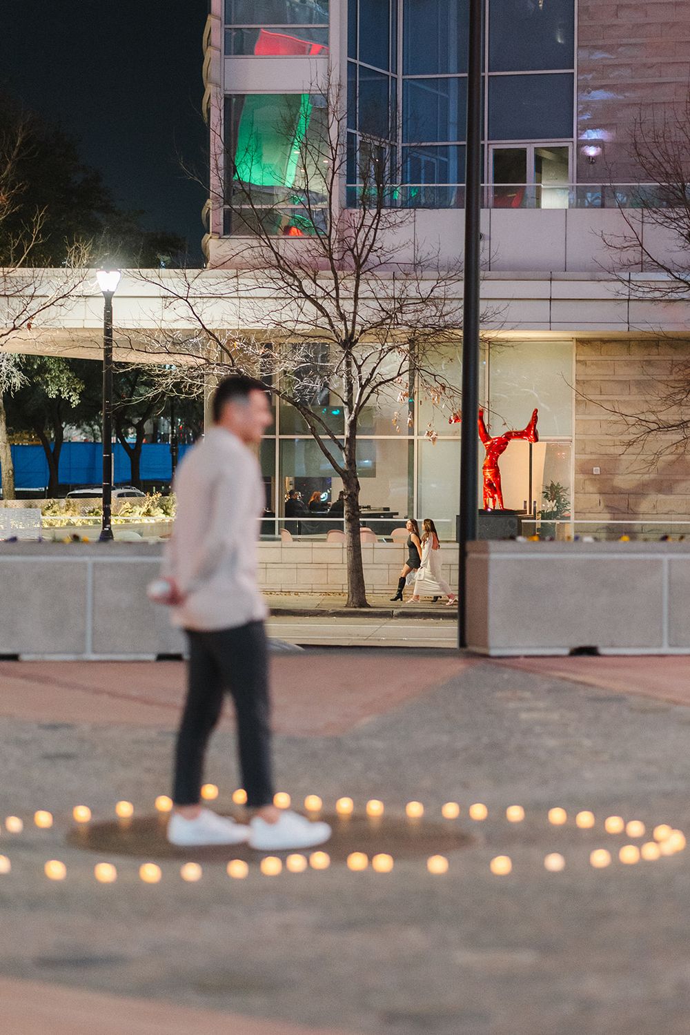 Person stands in a circle of candlelight outside a modern building with illuminated windows at night.