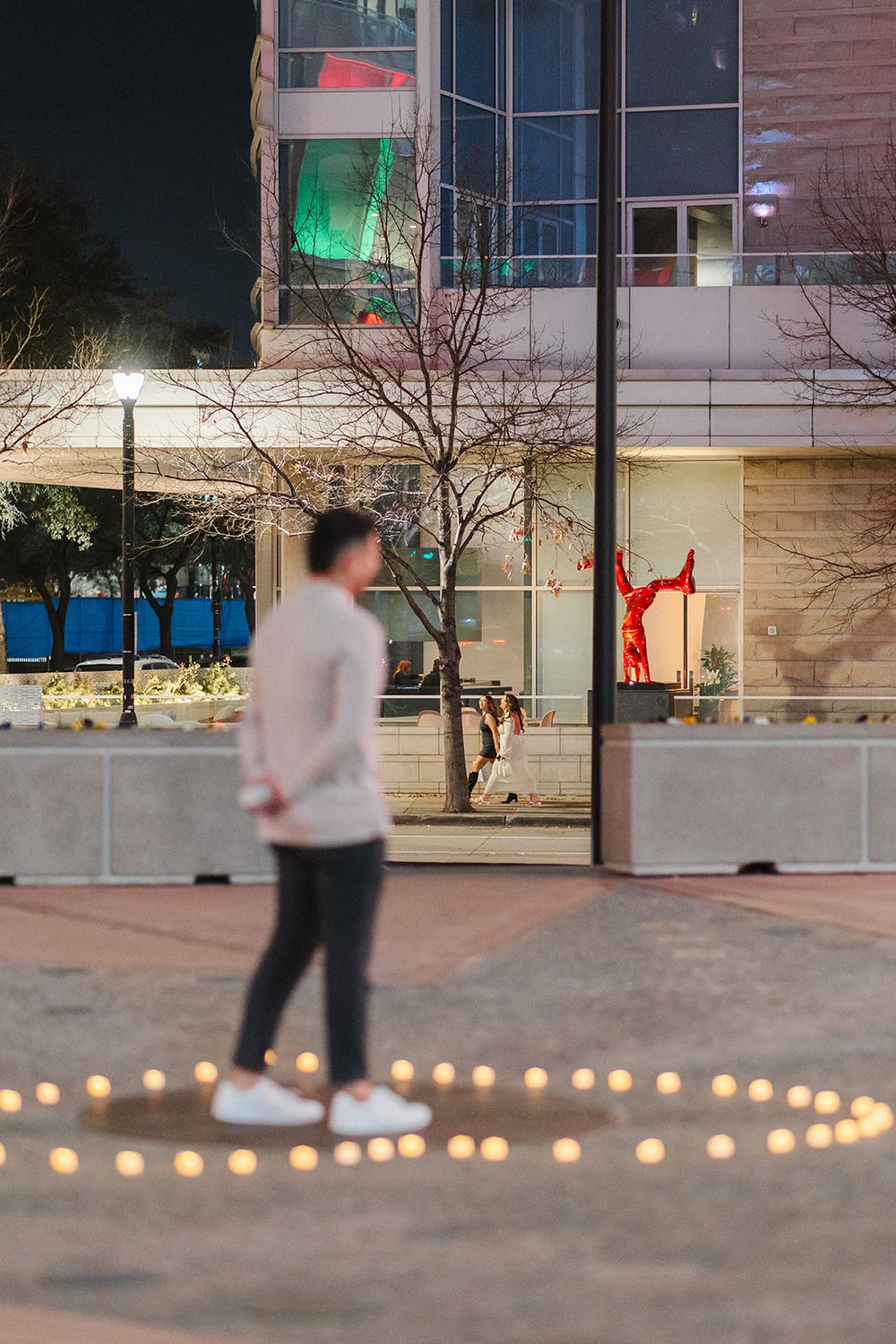 Someone stands on a plaza at night surrounded by glowing candles arranged in a heart shape on the ground.