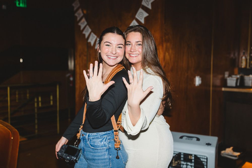 Two friends in casual outfits smile and wave at the camera while posing together in a dimly lit venue.