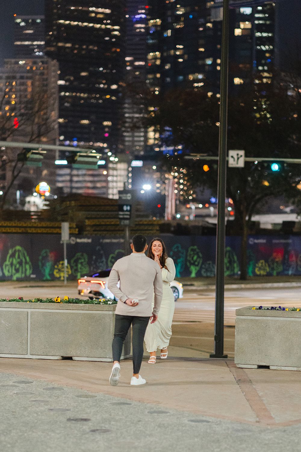 A couple stands together at night on a city plaza with illuminated skyscrapers and street art in the background.