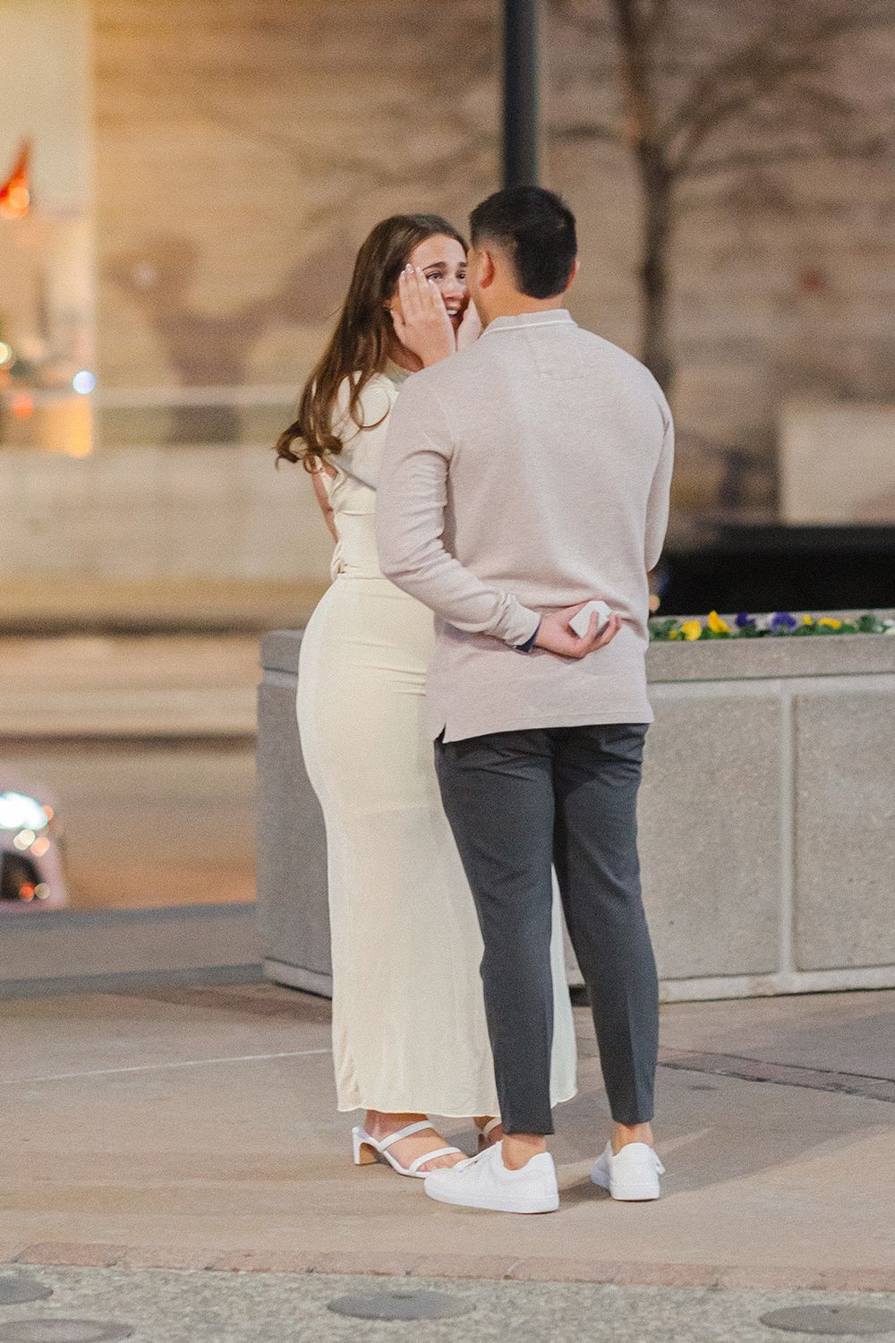 Couple embraces during golden hour outside, with person wearing white dress and another in casual attire with sneakers.
