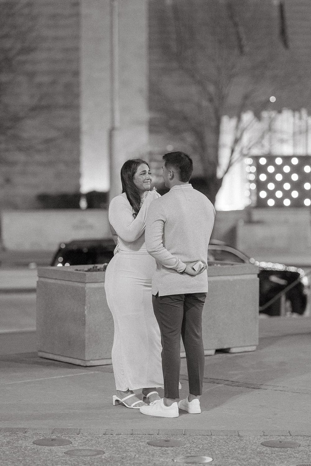 A couple embraces during a nighttime proposal moment with decorative lights in the background.