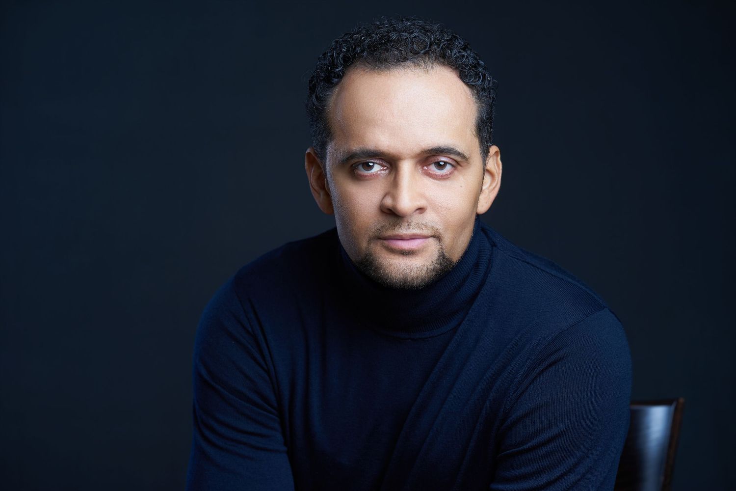 Man wearing a navy turtleneck sweater poses for a professional portrait against a dark background.