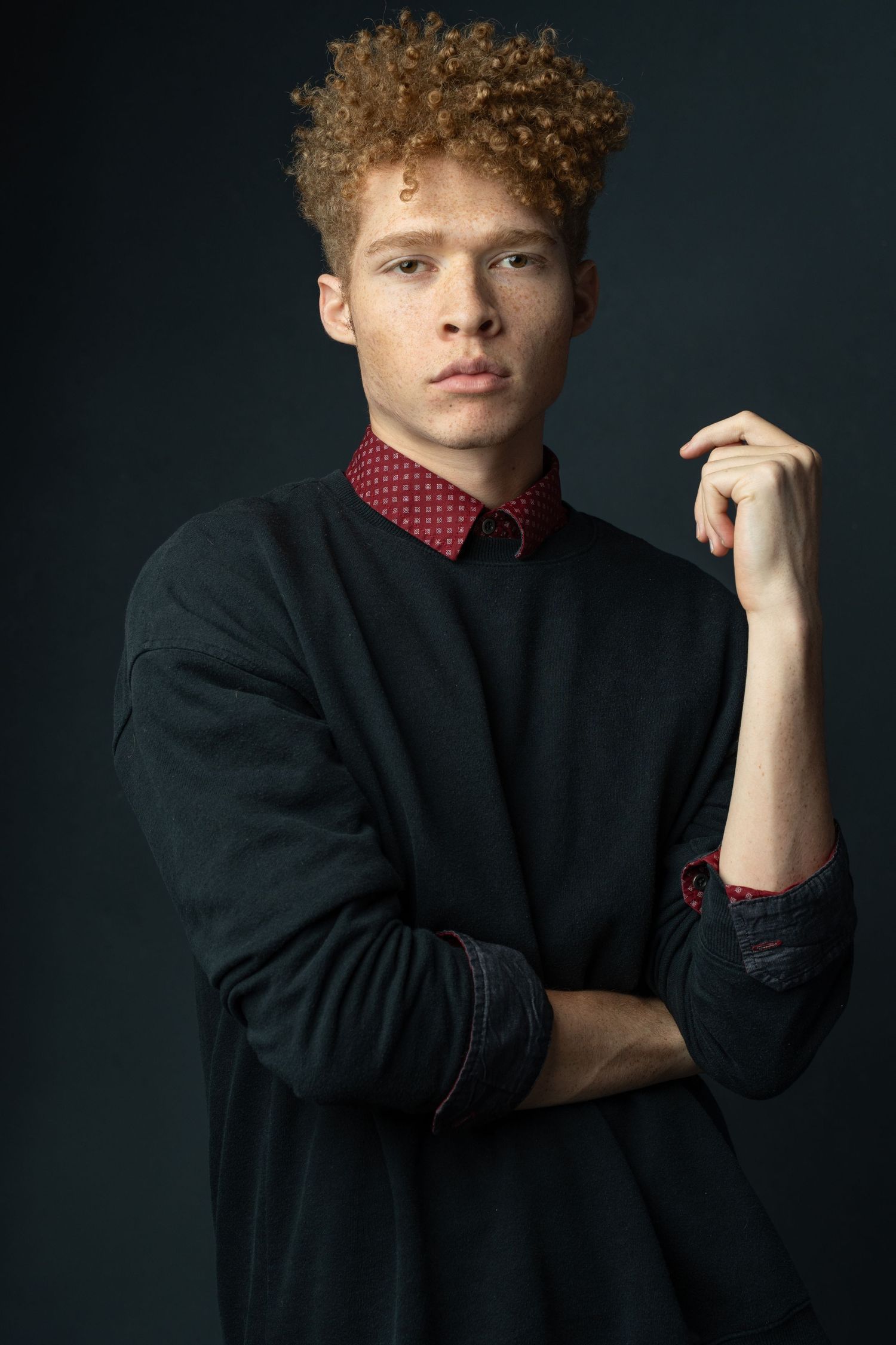 Young man with curly red hair wearing a black sweater over a red collared shirt poses against dark background.