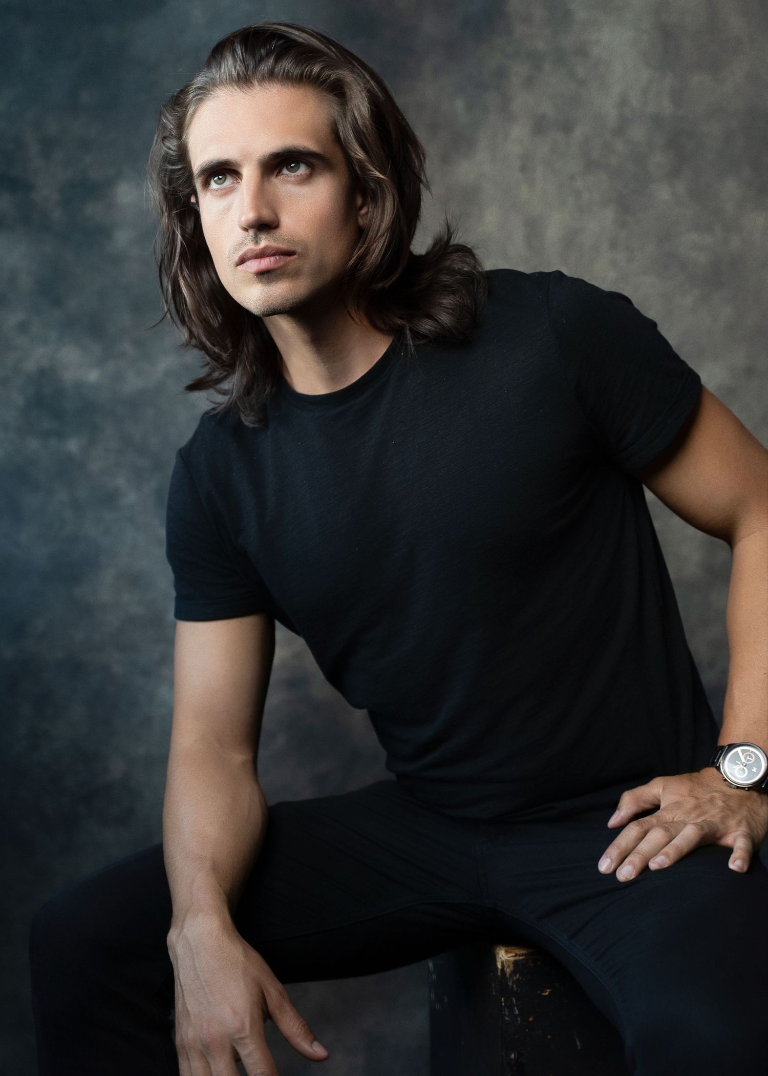 Young man with long brown hair wearing a black t-shirt sits confidently against a gray studio backdrop.