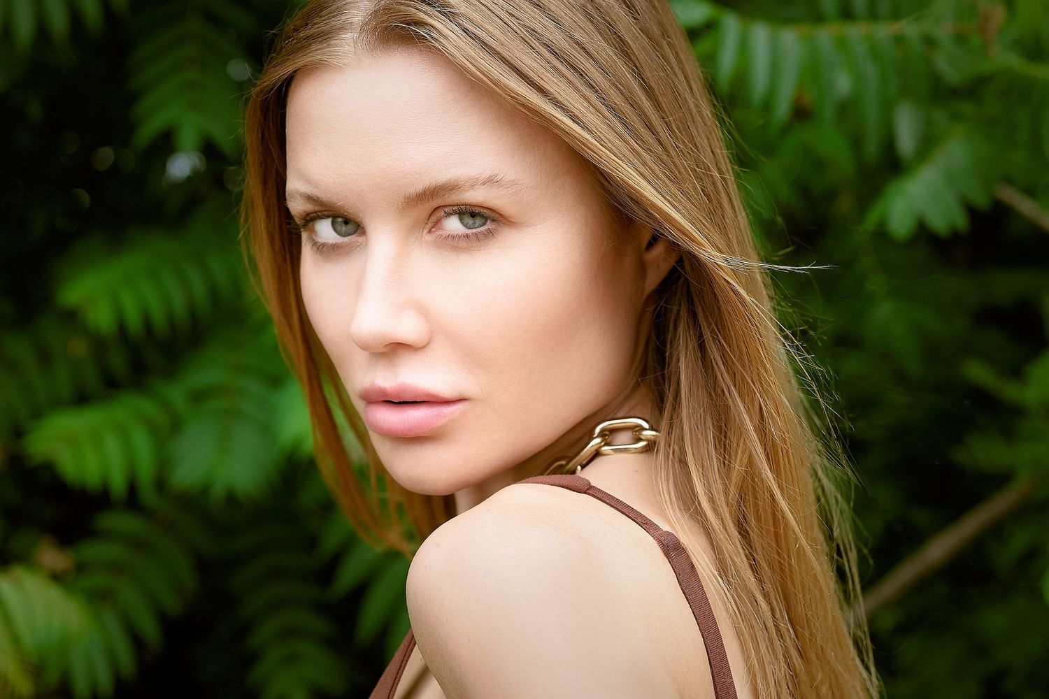 Blonde woman with straight hair in a brown strap top poses outdoors near lush green foliage.