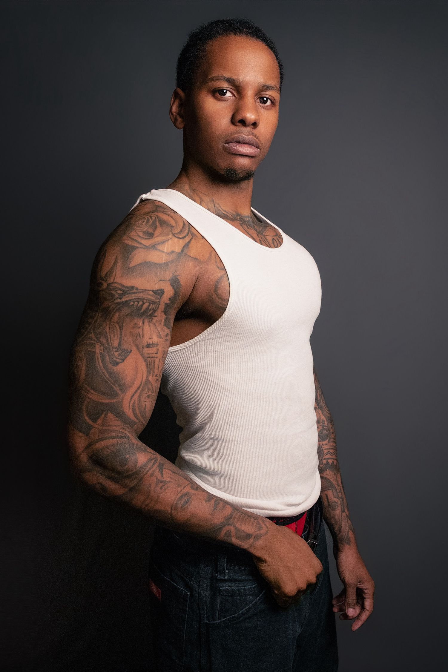 Tattooed man in a white tank top poses against a dark background showcasing sleeve tattoos.