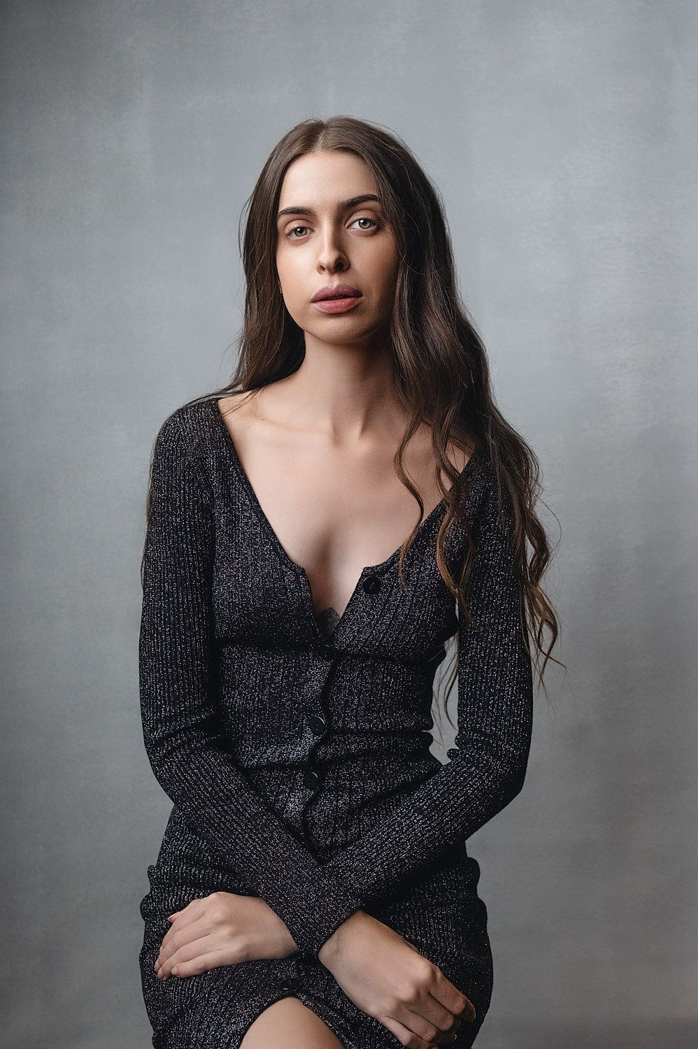 Brunette woman in a dark knit low-cut dress poses seated against a gray studio background.