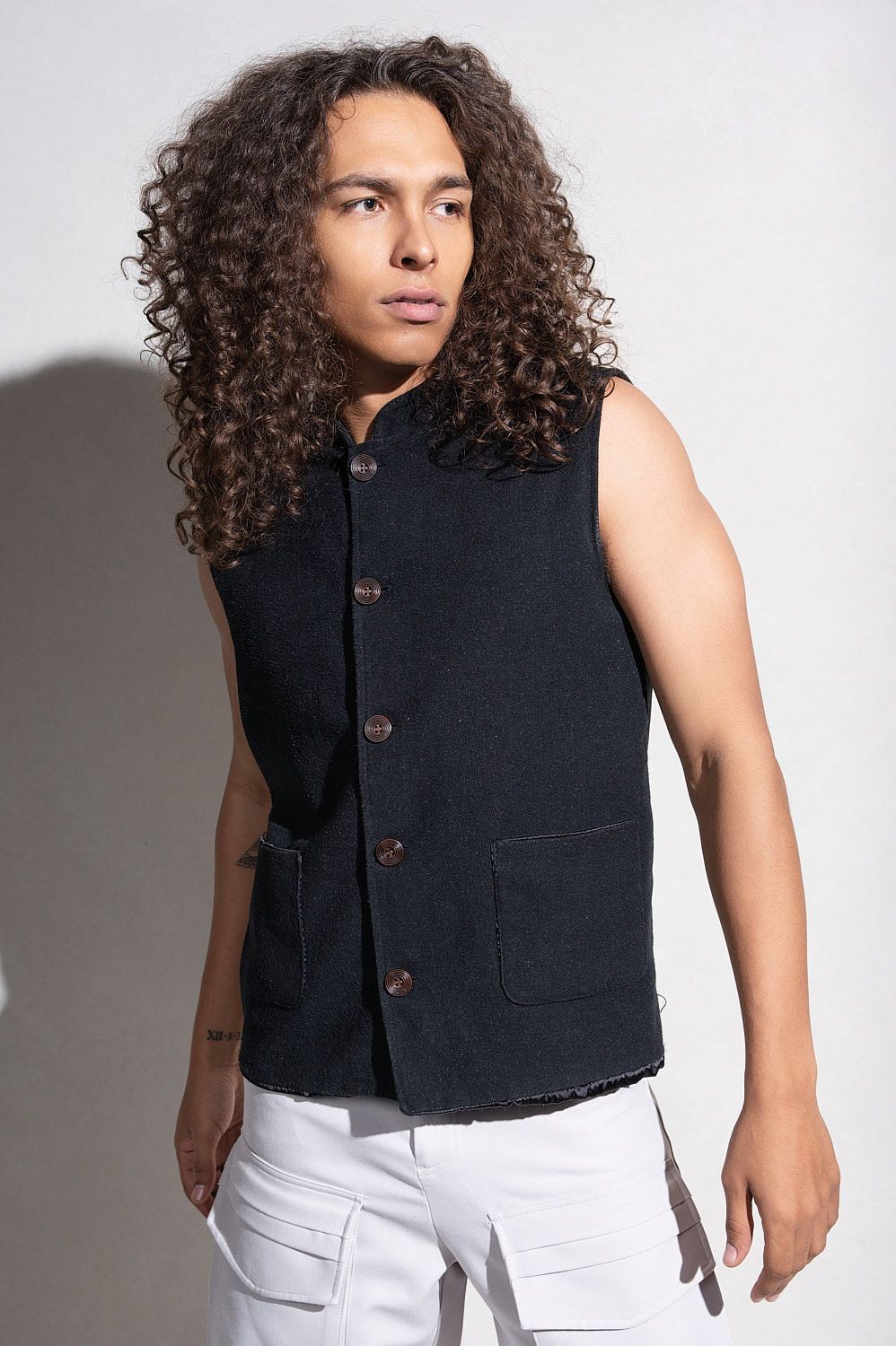 Young man with long curly hair wearing a dark sleeveless denim vest and white cargo pants.