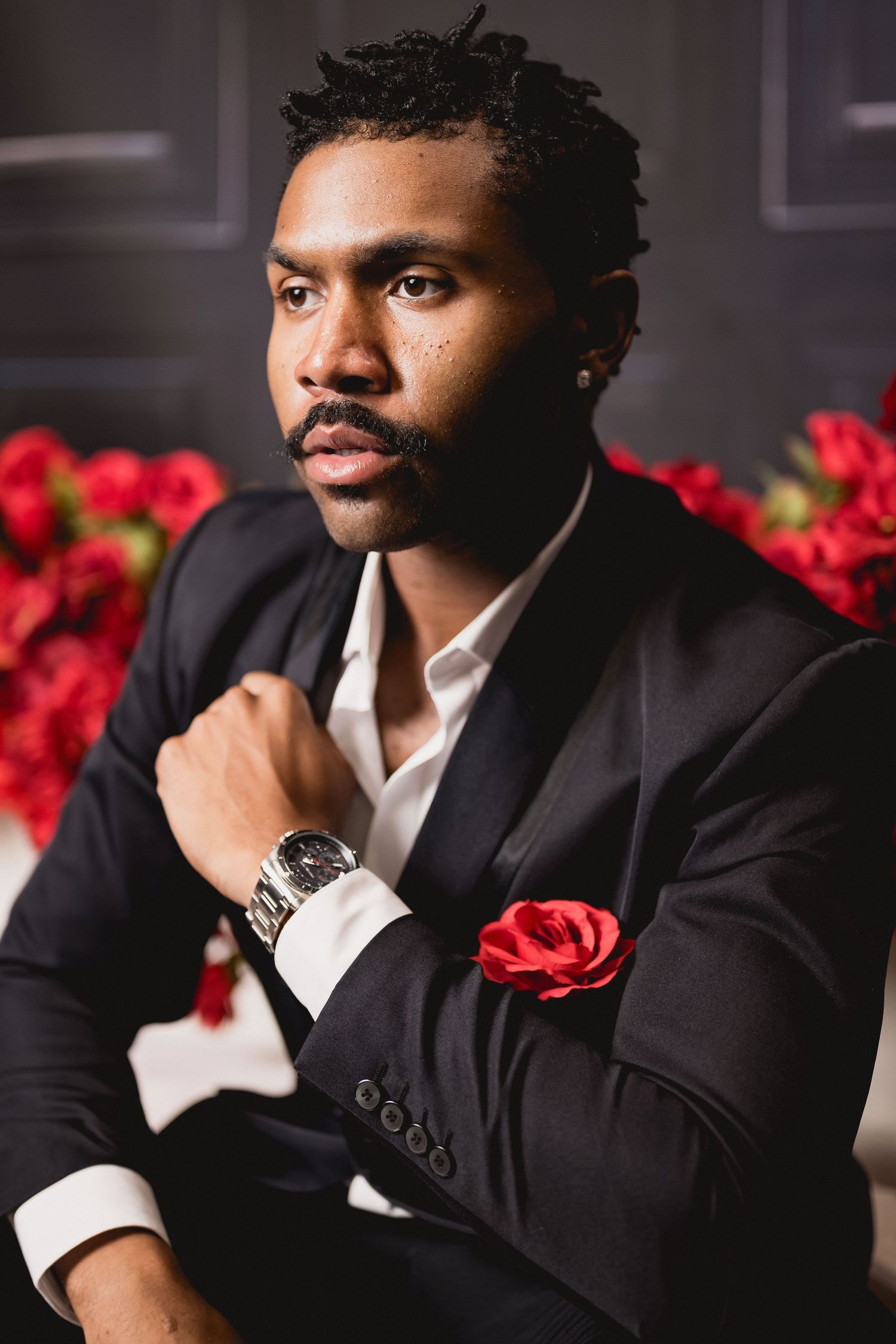 Stylish man in a black suit with a red pocket square and watch poses near red roses in a dark setting.
